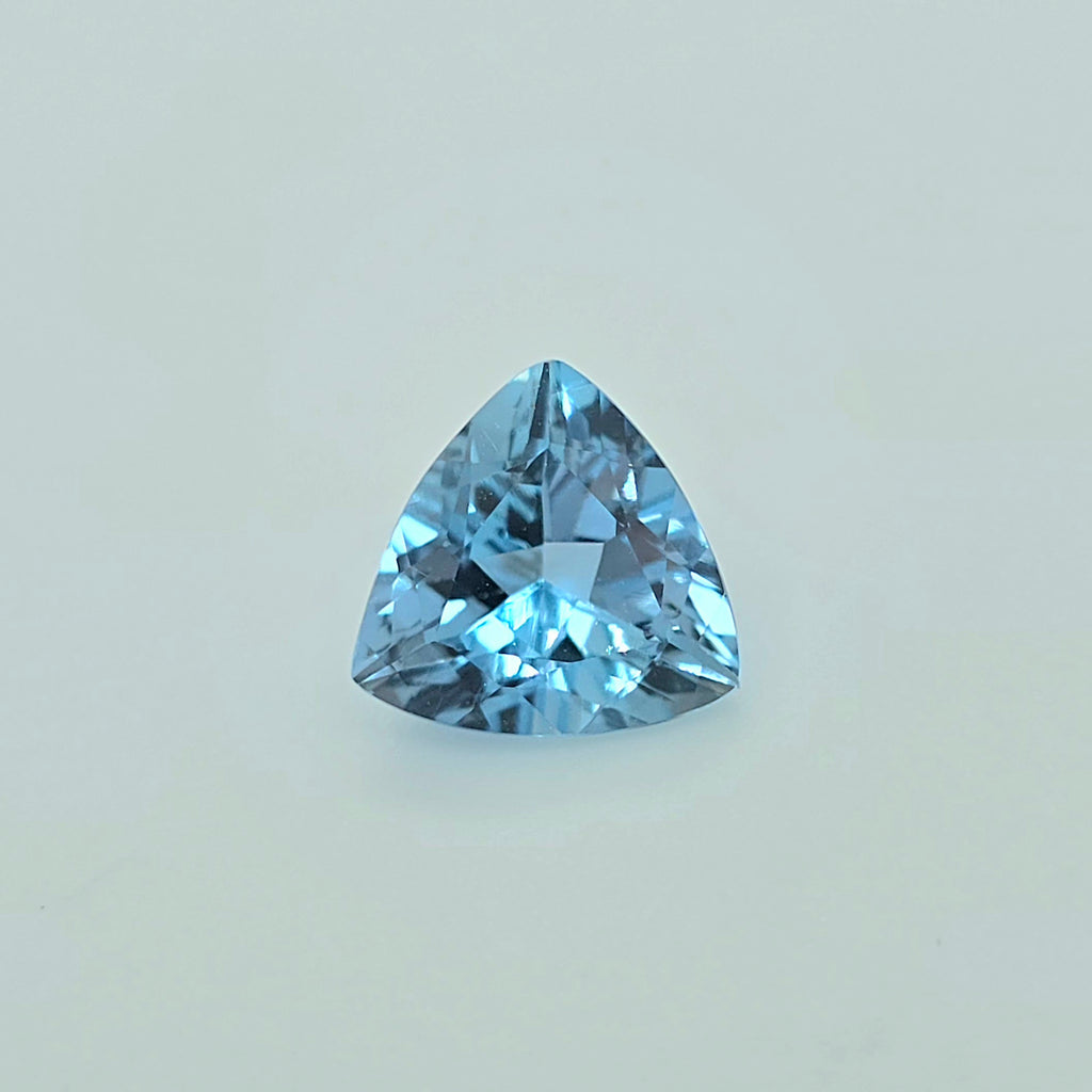A single triangular light blue gemstone, faceted and cut to sparkle, centered on a clean, light off-white background.