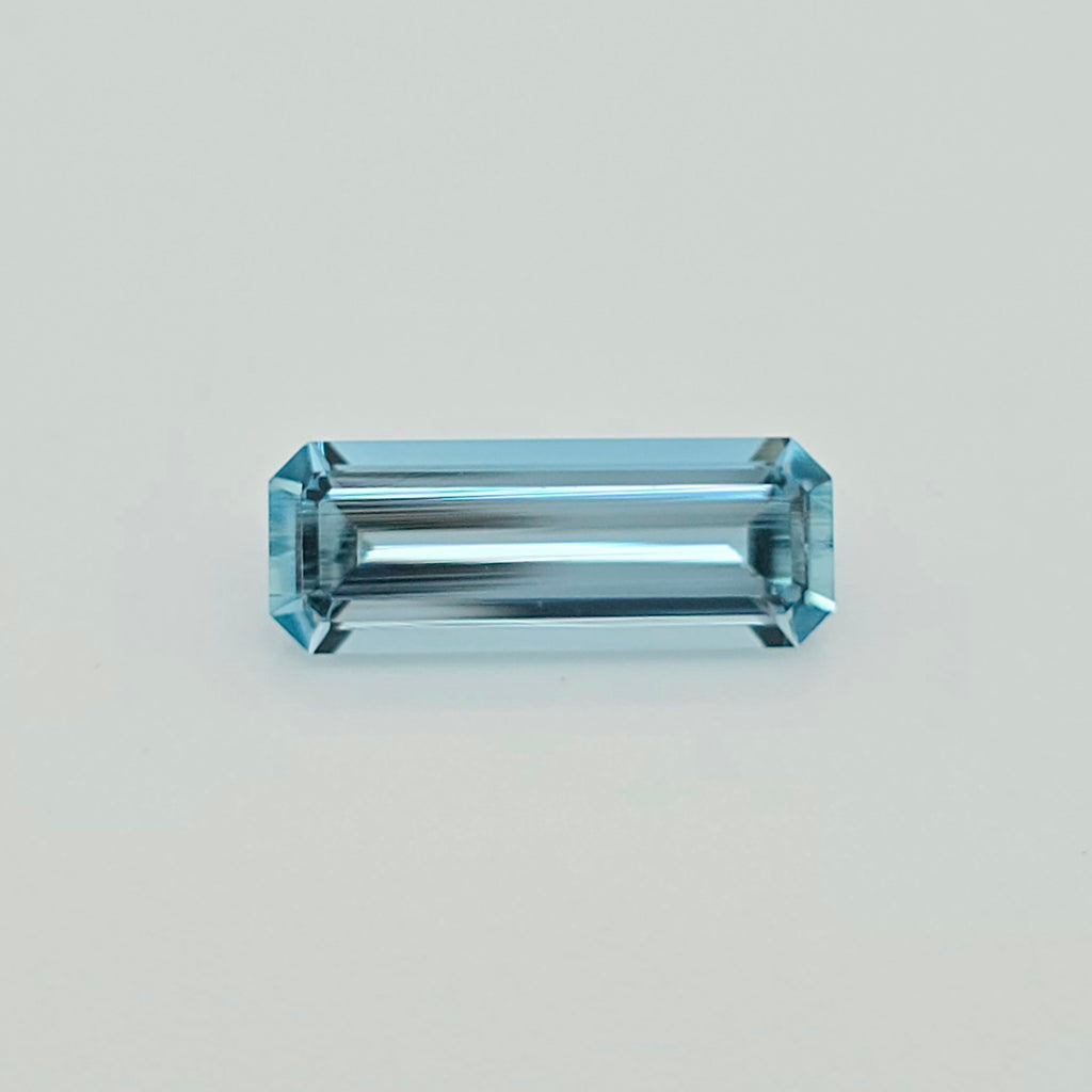A rectangular emerald-cut light blue gemstone with a faceted, polished finish, centered on a clean, light gray background.