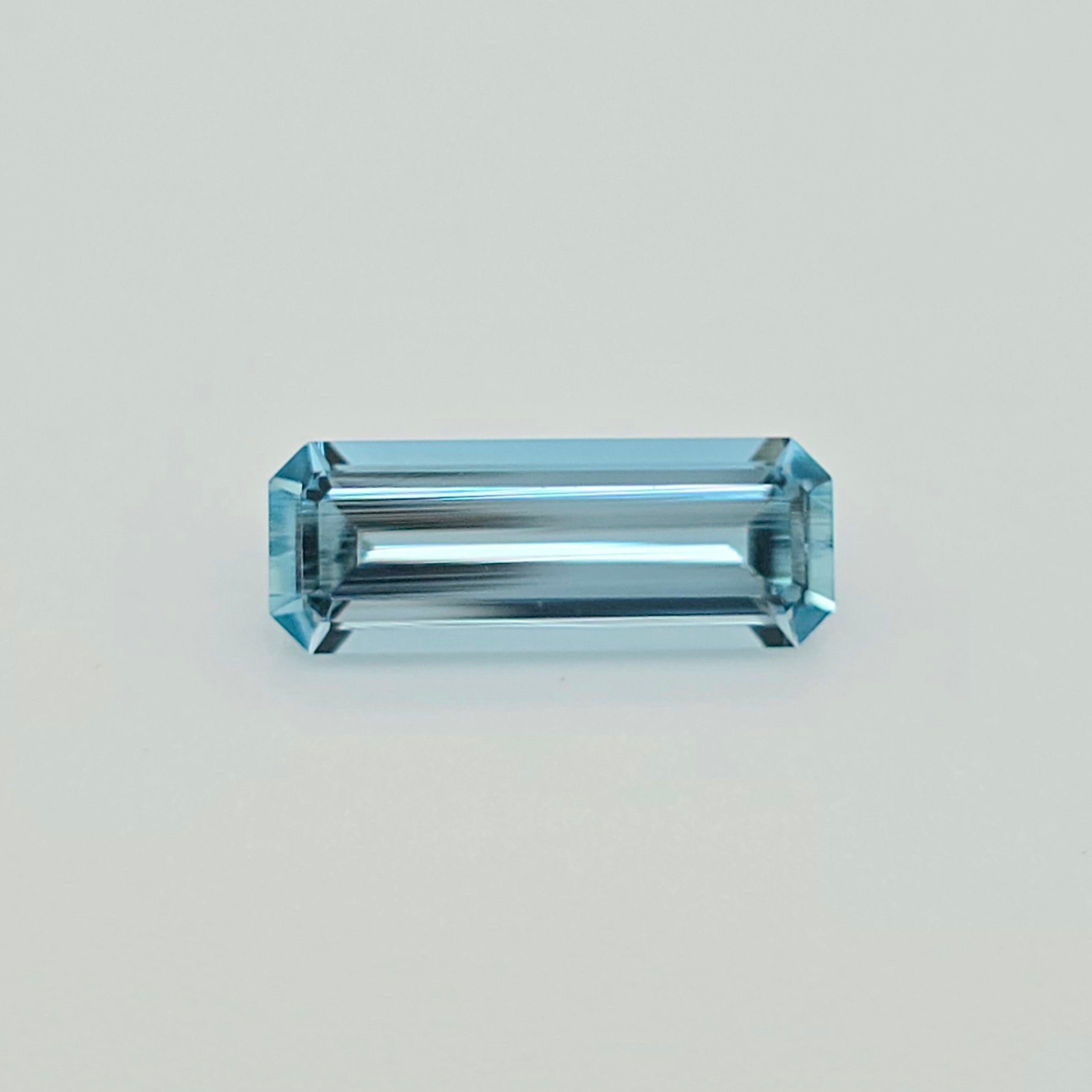 A rectangular emerald-cut light blue gemstone with a faceted, polished finish, centered on a clean, light gray background.