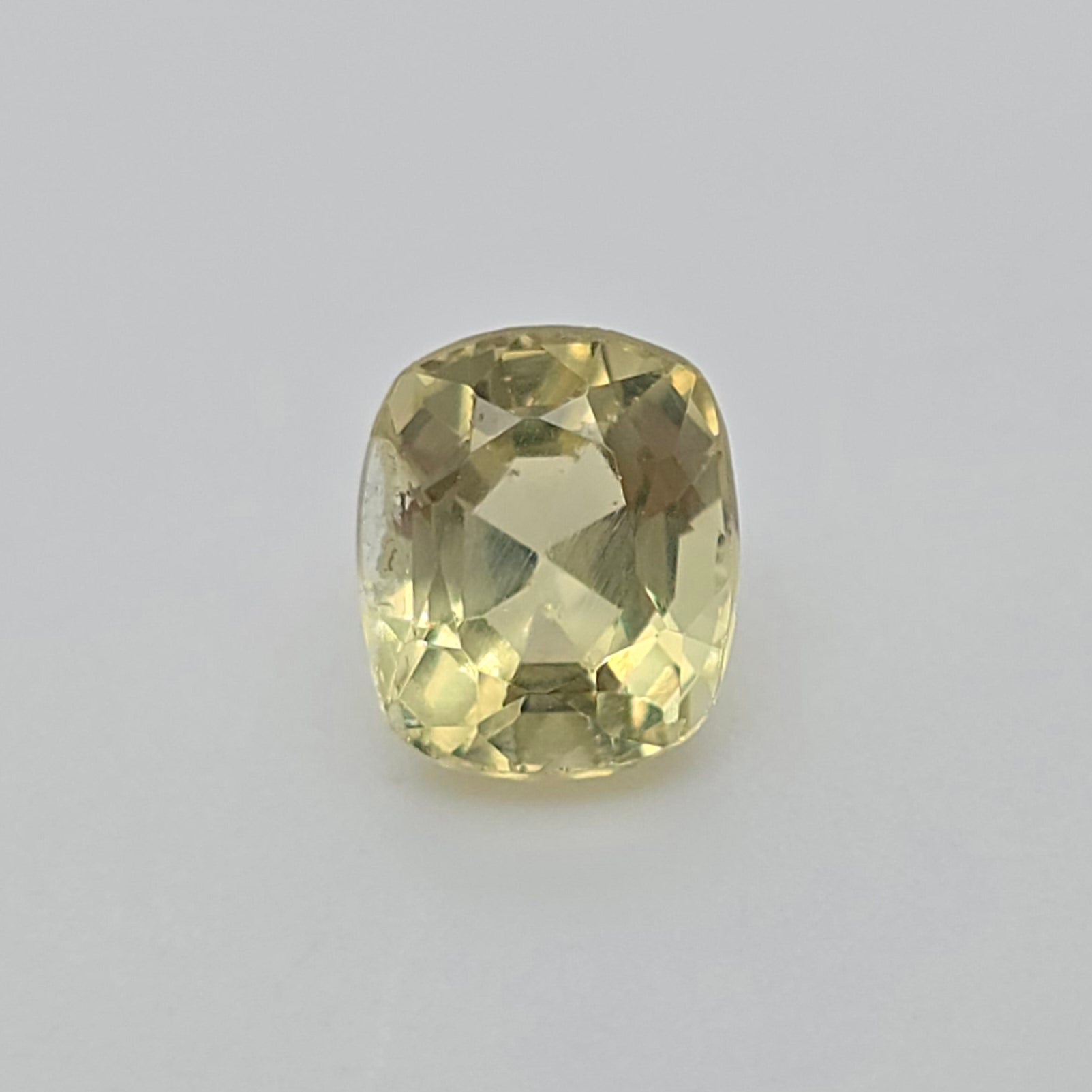 A cushion-cut yellow gemstone with a faceted cushion shape, displayed on a neutral light gray background.