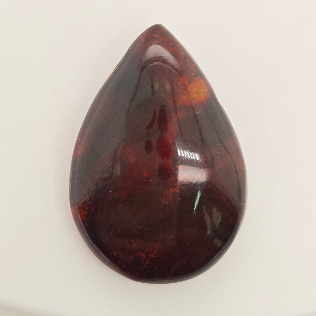 A polished teardrop-shaped gemstone with a deep reddish-brown color and natural inclusions, shown on a plain off-white background.