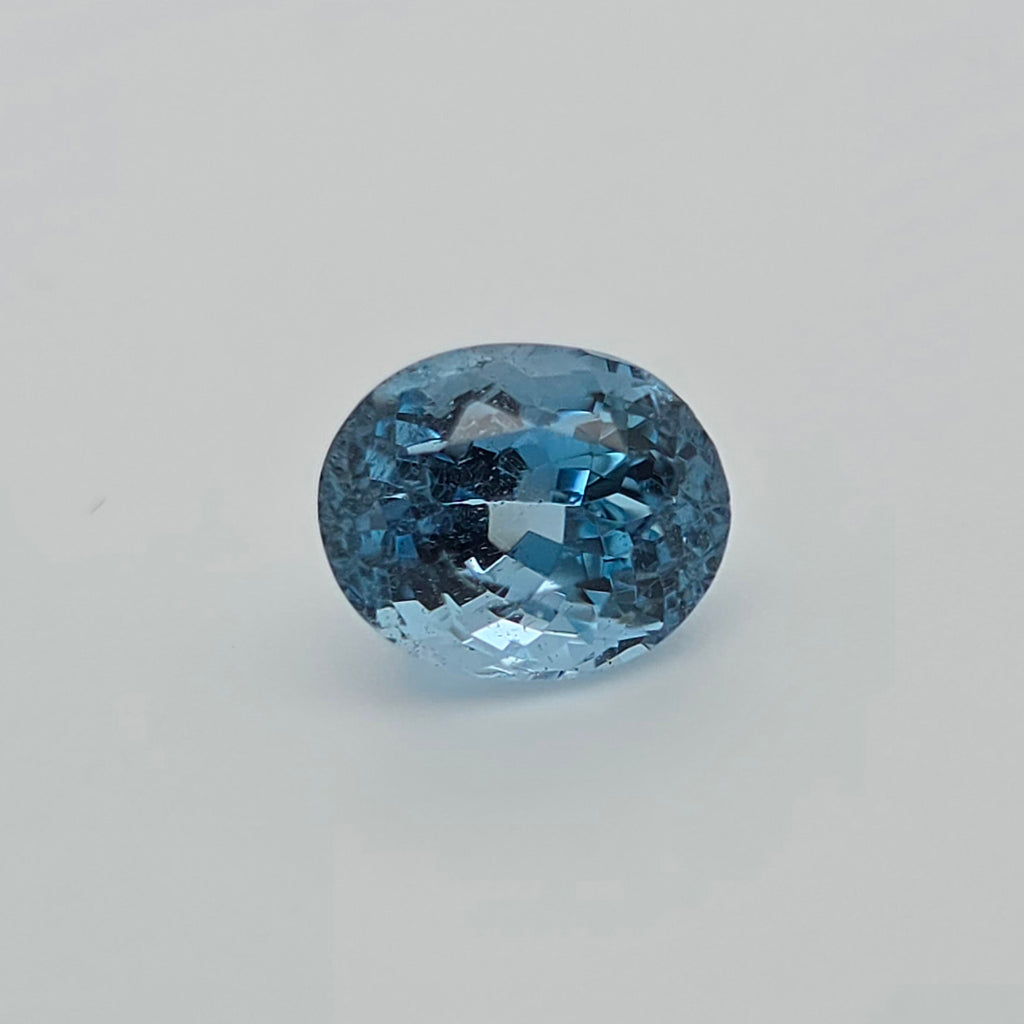 An oval-cut light blue gemstone with a faceted, sparkling surface, centered on a plain light gray background.