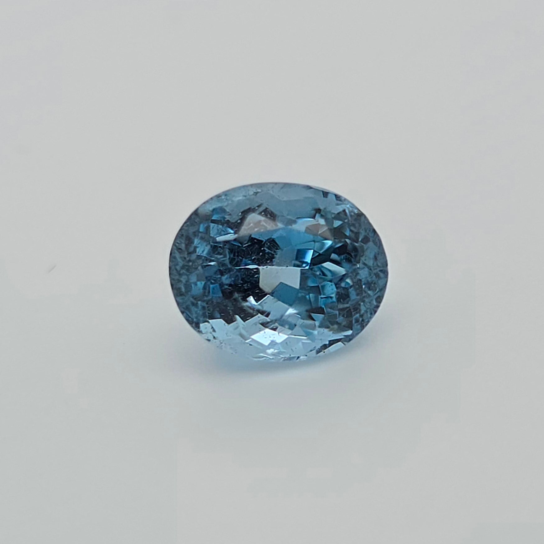 An oval-cut light blue gemstone with a faceted, sparkling surface, centered on a plain light gray background.