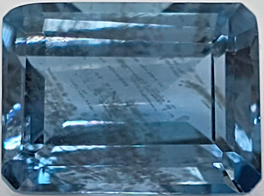 Close up image of a faceted emerald cut blue gemstone with natural inclusions on a plain white background