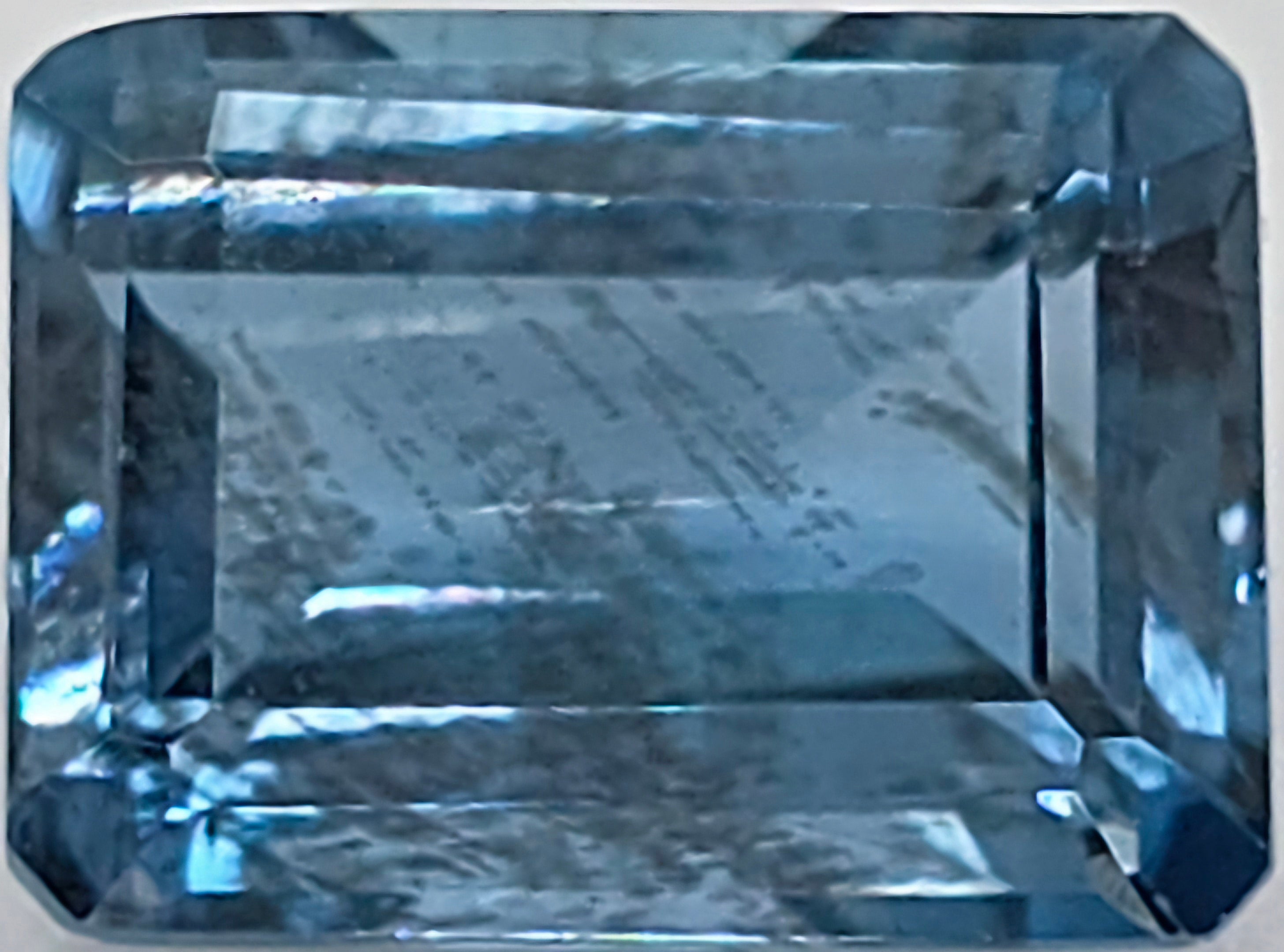Close up image of a faceted emerald cut blue gemstone with natural inclusions on a plain white background