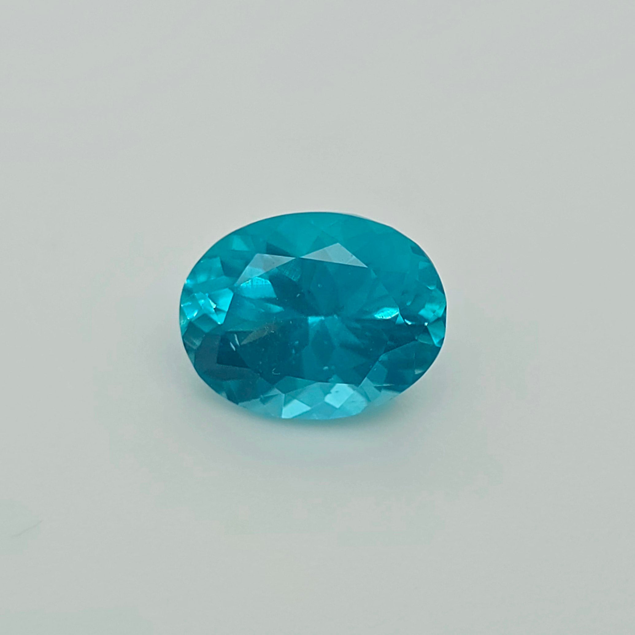 An oval-cut, faceted turquoise gemstone on a plain light gray background.
