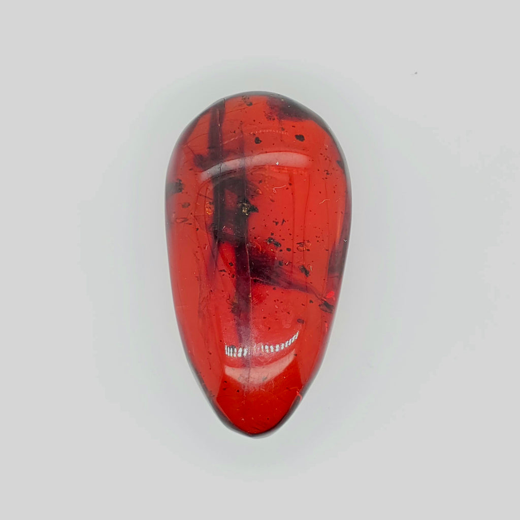 A polished teardrop-shaped reddish orange gemstone with dark inclusions and speckles, displayed on a light gray background.