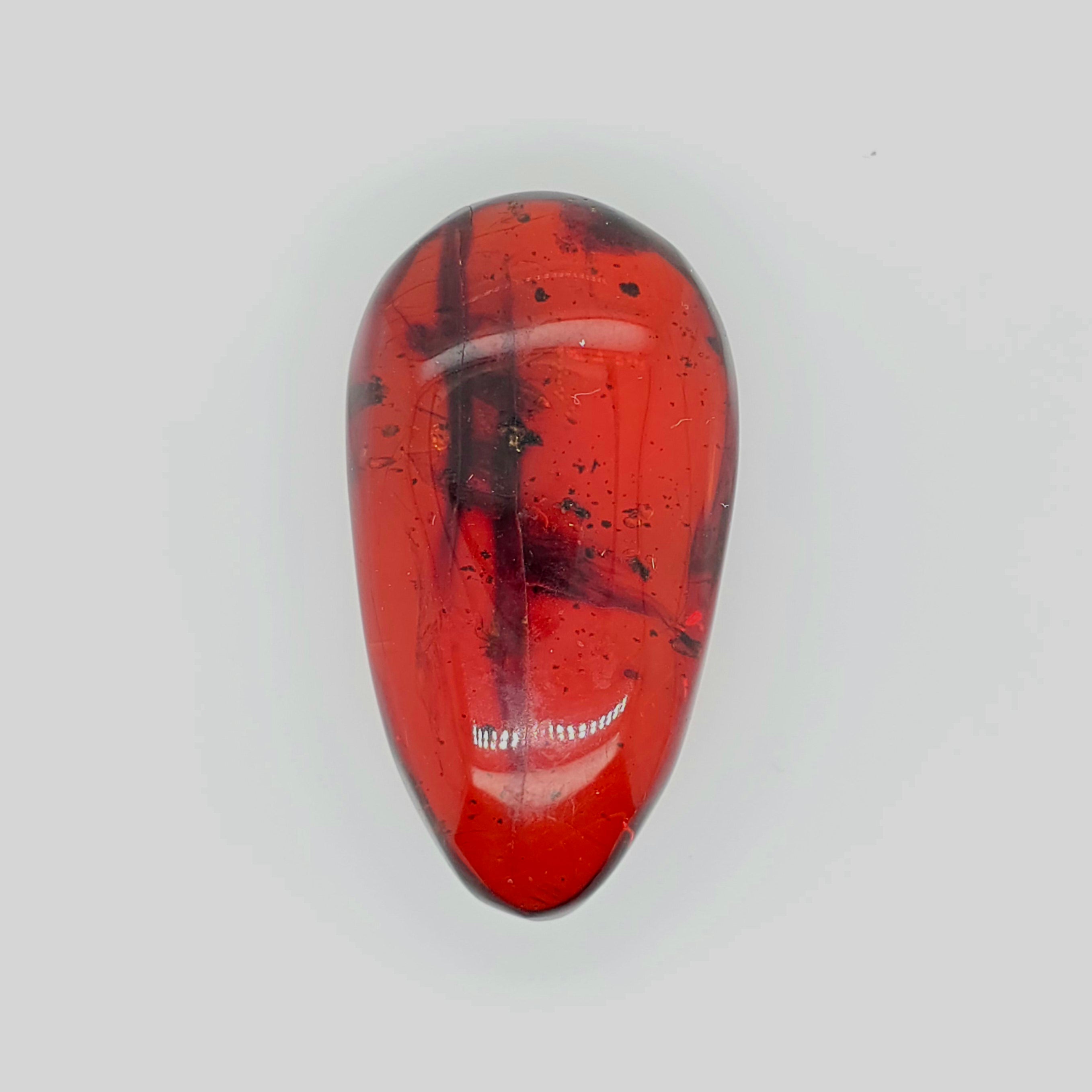 A polished teardrop-shaped reddish orange gemstone with dark inclusions and speckles, displayed on a light gray background.