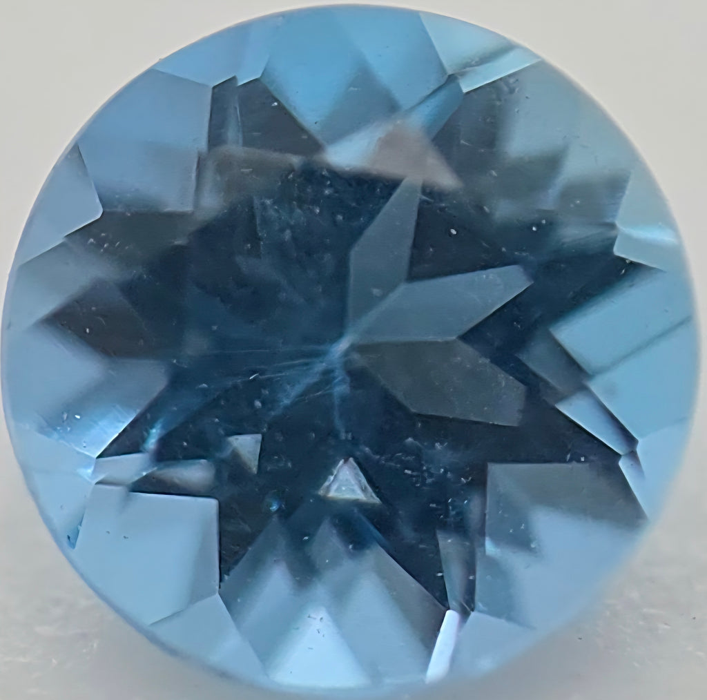 A round, faceted blue gemstone, shown in a close-up studio shot against a neutral light gray background.