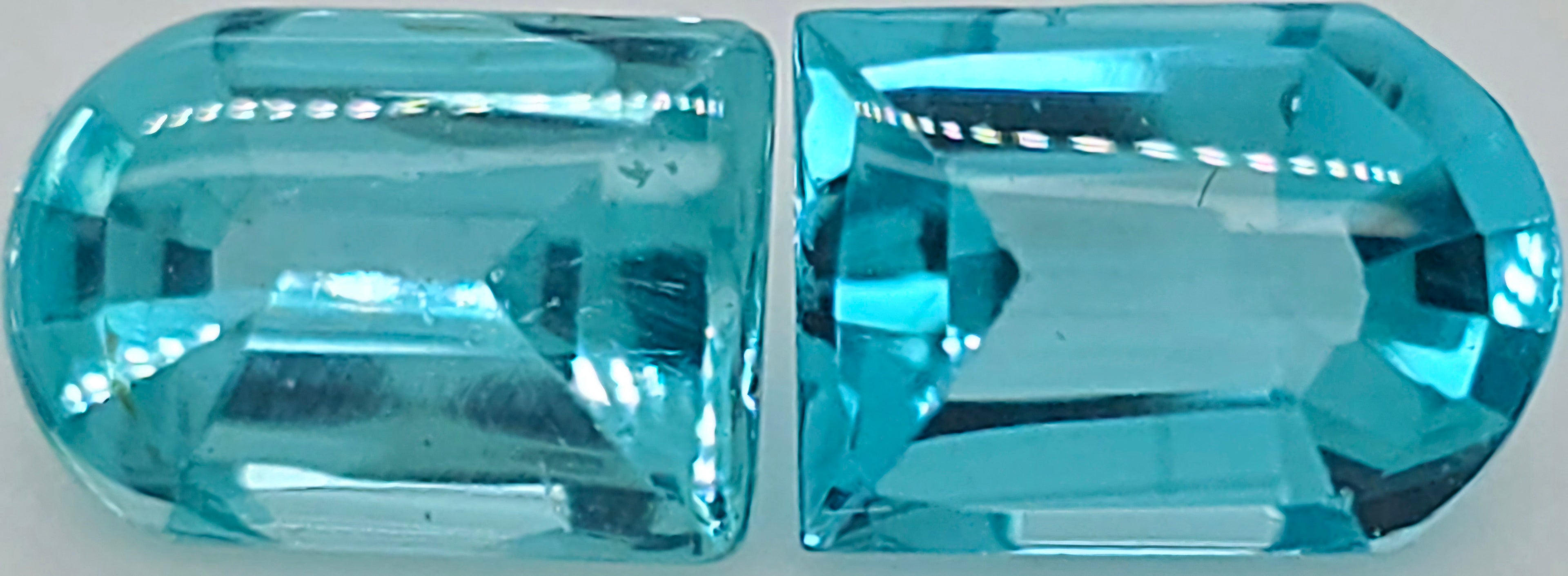 Two rounded rectangular cut greenish blue gemstones displayed side by side on a plain white surface.