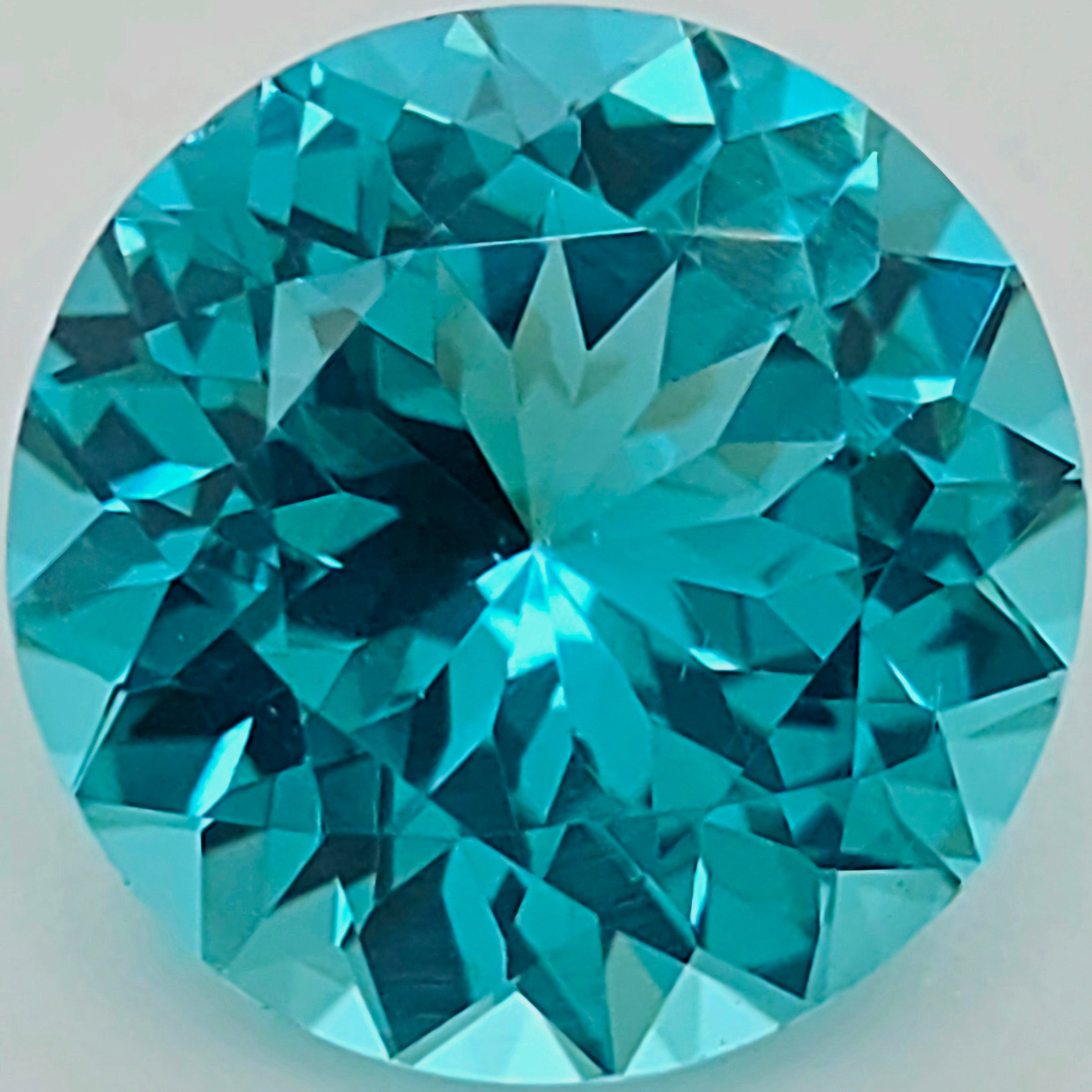 A round brilliant-cut greenish blue gemstone on a light gray background.