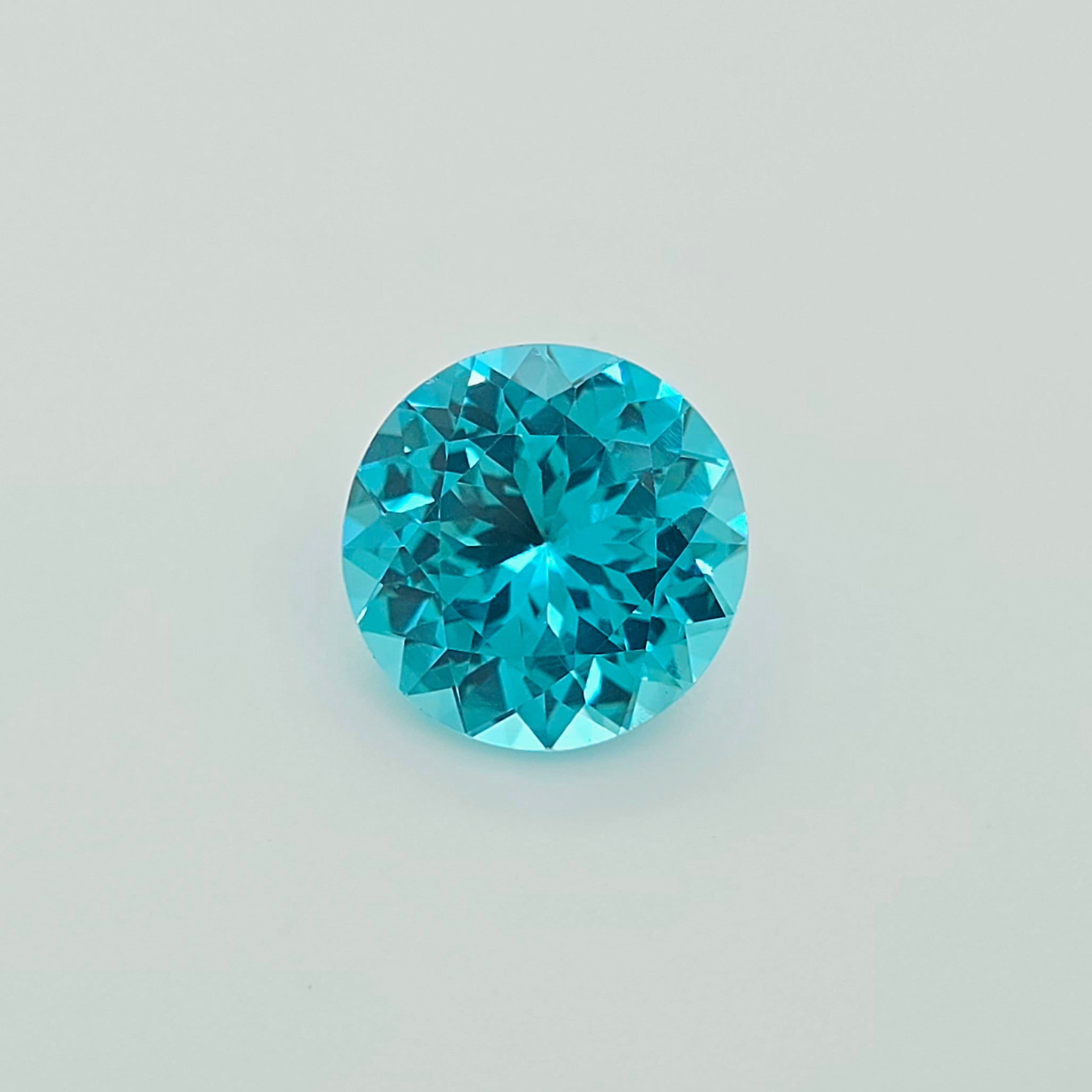 A round brilliant-cut greenish blue gemstone, centered on a clean, light gray background.