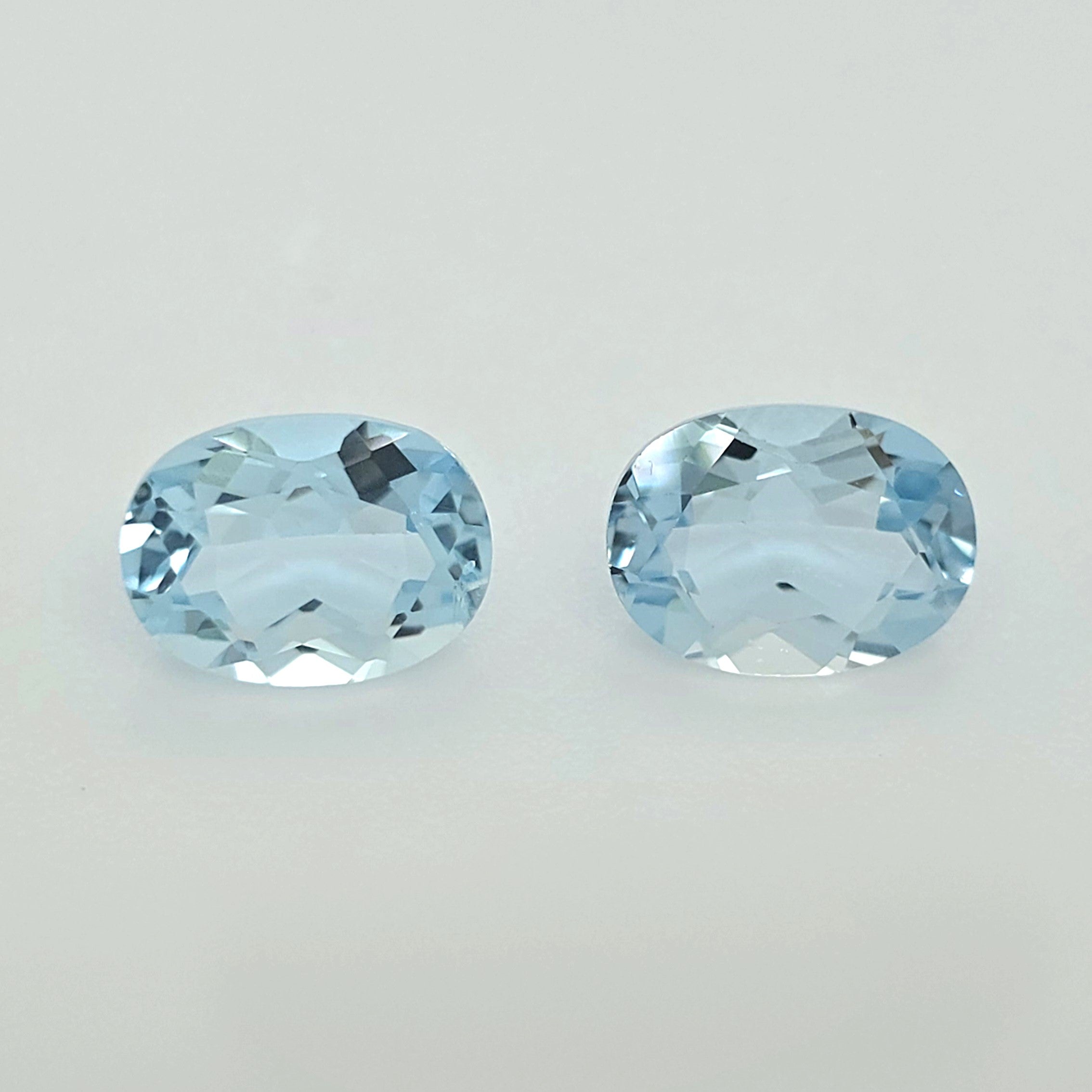 Two oval-cut light blue gemstones with faceted cuts, displayed side-by-side on a plain light gray background.