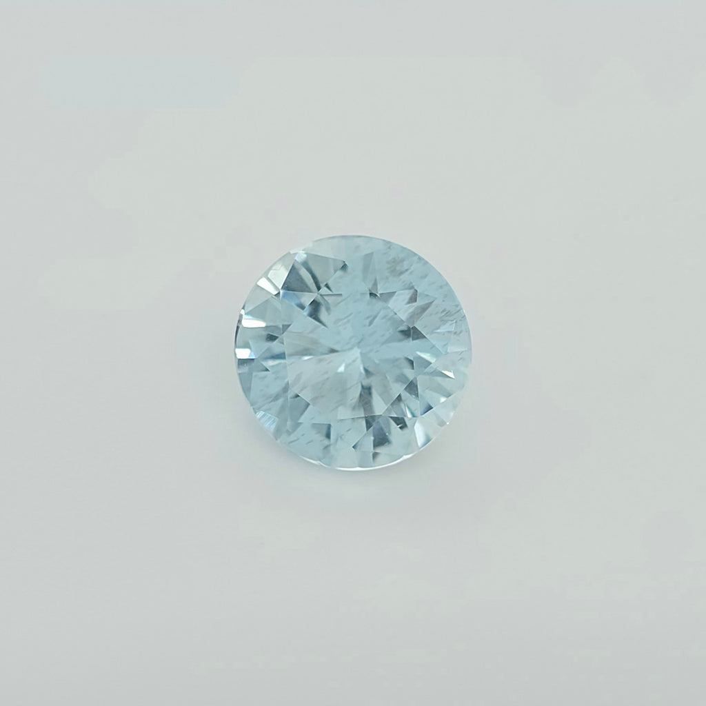 A round brilliant-cut light blue gemstone, centered on a clean, light gray background.