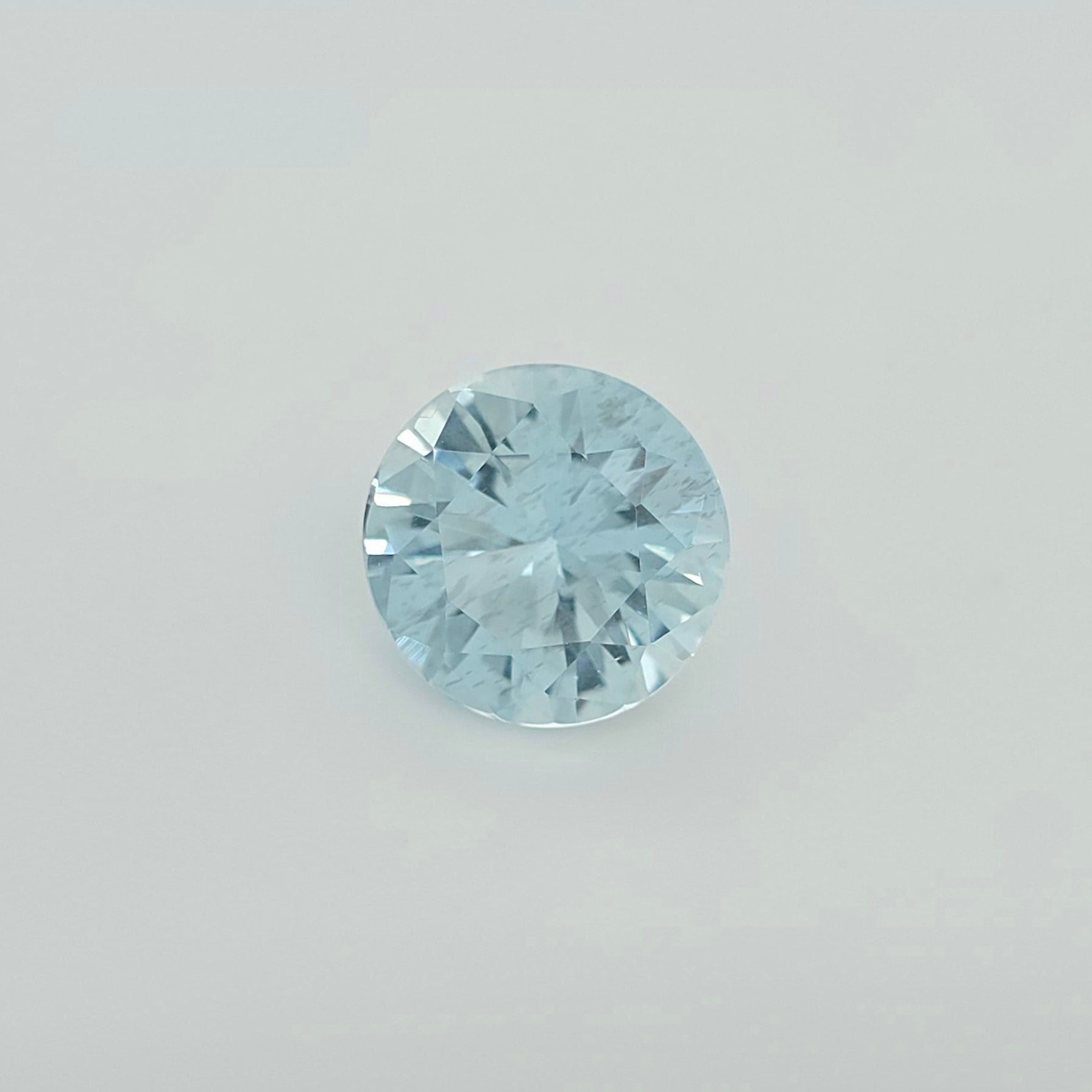 A round brilliant-cut light blue gemstone, centered on a clean, light gray background.