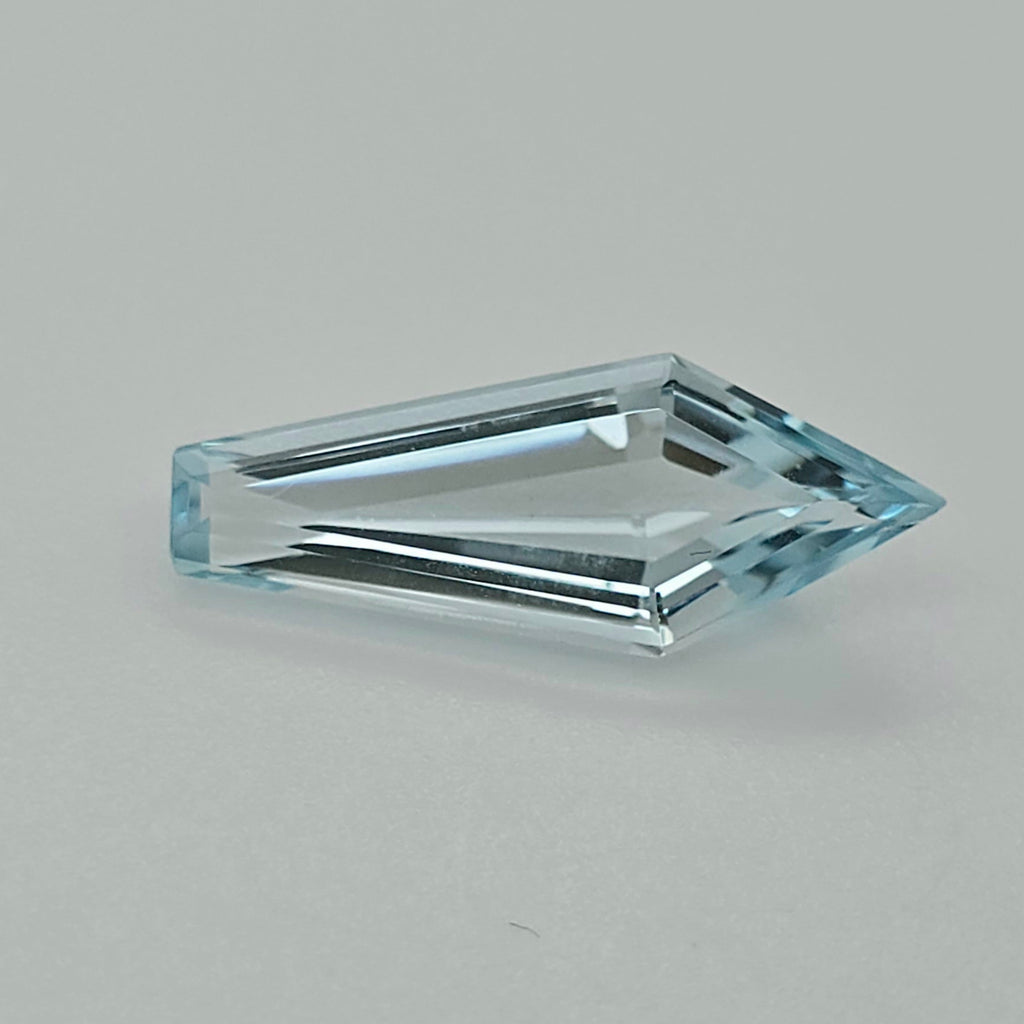 A single faceted light blue gemstone with an elongated kite shape, shown on a neutral light gray background.