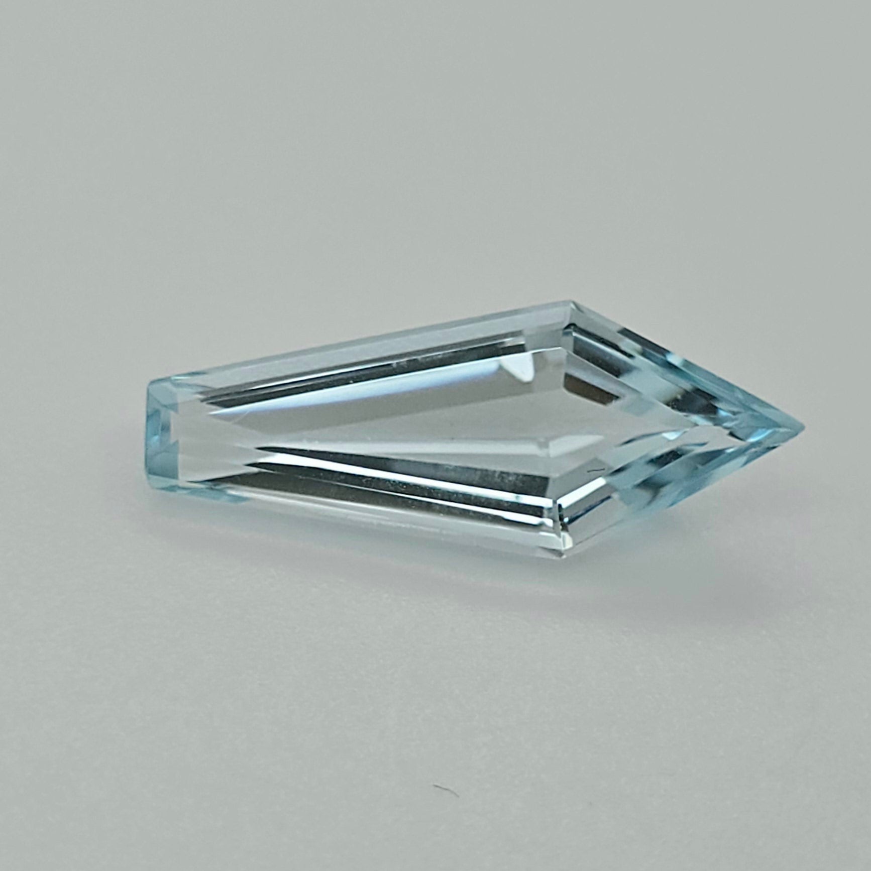 A single faceted light blue gemstone with an elongated kite shape, shown on a neutral light gray background.