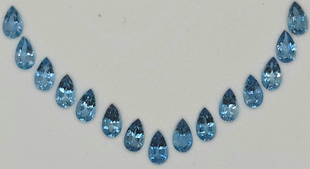 A row of 15 faceted pear-cut blue gemstones arranged in a curved line on a plain white background.