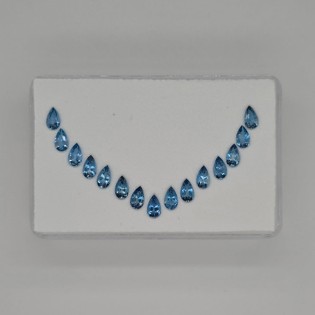 A set of 15 teardrop-shaped blue gemstones arranged in a curved row on a white backing card, photographed against a neutral gray background.