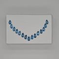A set of 15 teardrop-shaped blue gemstones arranged in a curved row on a white backing card, photographed against a neutral gray background.