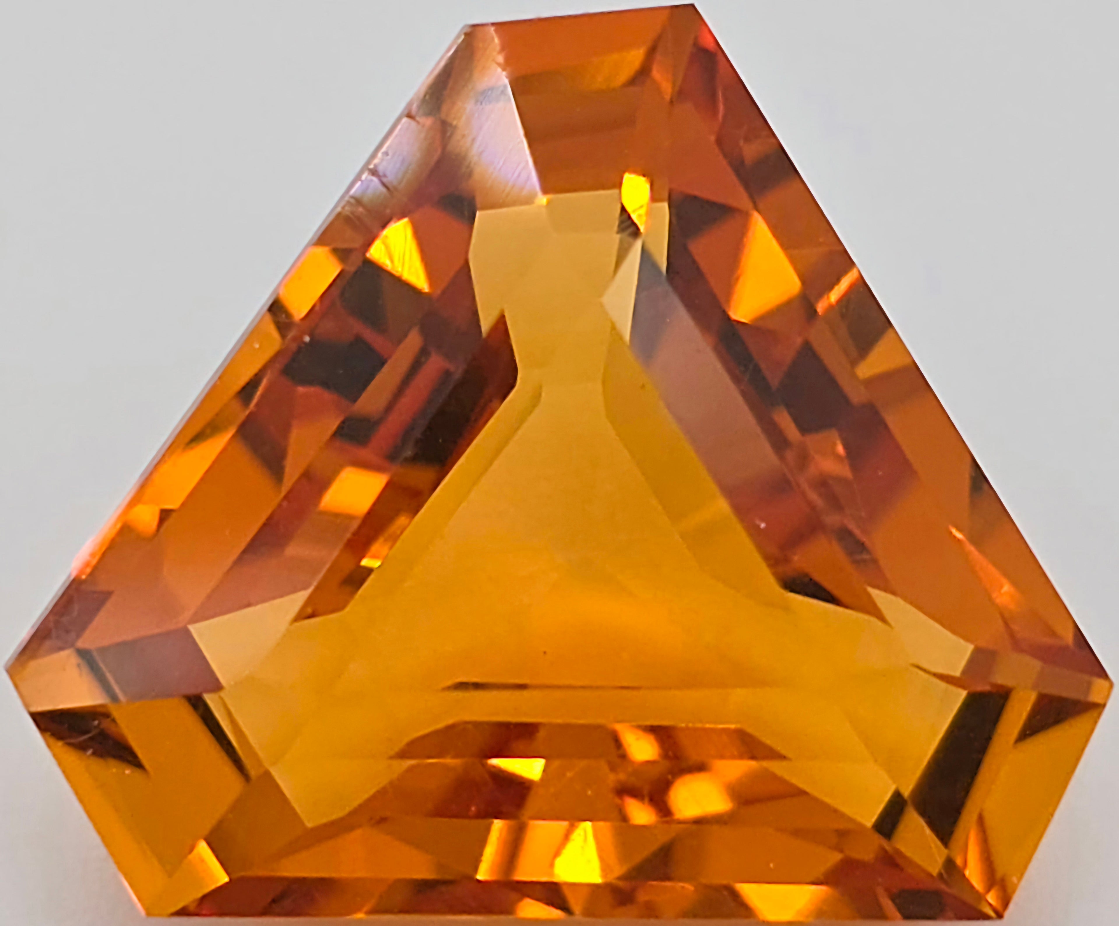 A faceted triangular gemstone in a vibrant orange color, shown in close-up against a neutral light gray background.