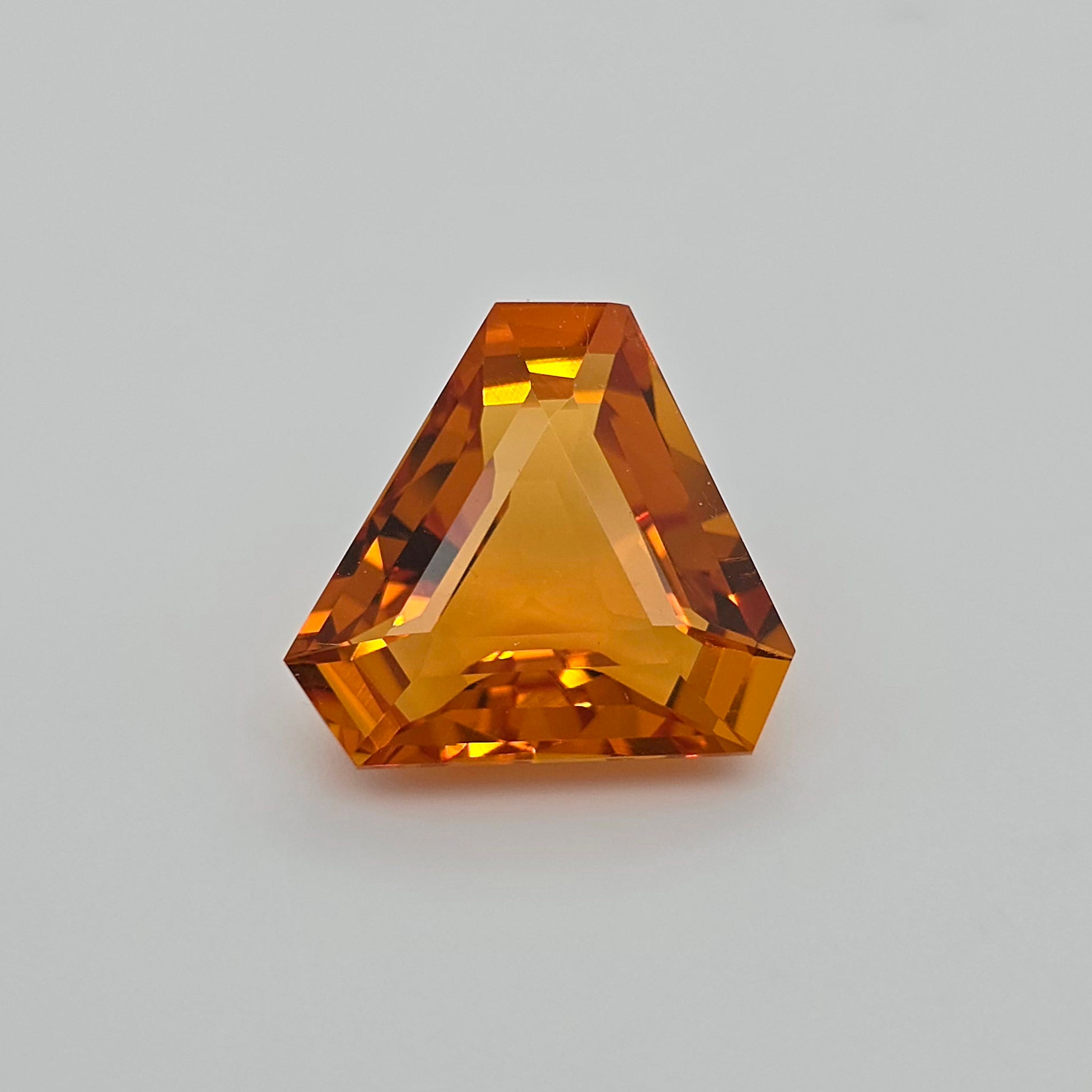 A faceted triangular orange gemstone, centered on a neutral light gray background.
