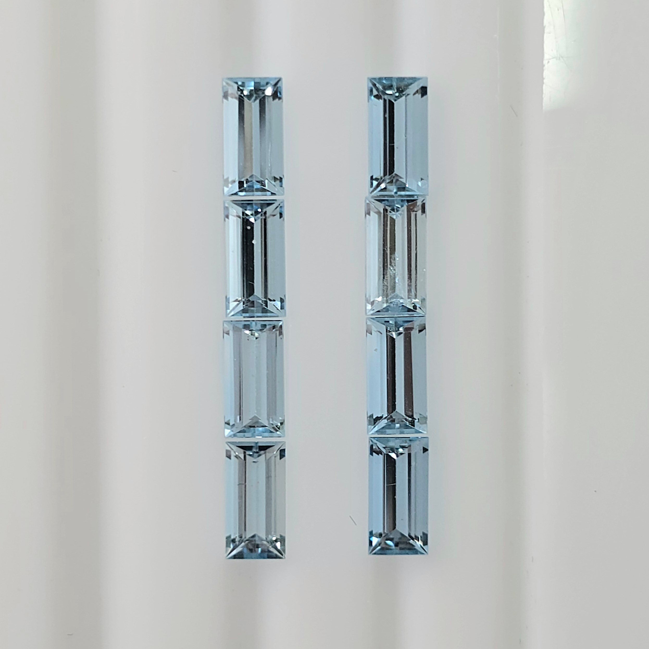 Eight baguette cut faceted light blue gemstones in two columns shown vertically on a plain light background.
