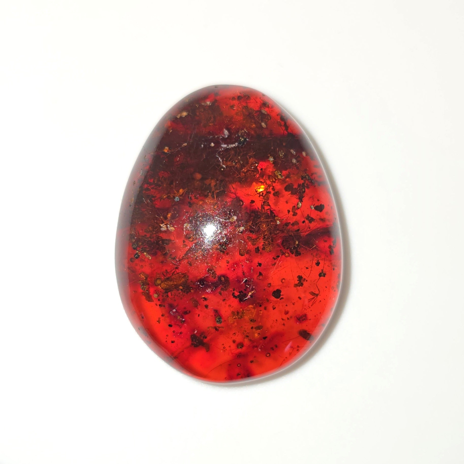 A polished red-orange cabochon gemstone with dark brown and black inclusions, photographed on a plain white background.