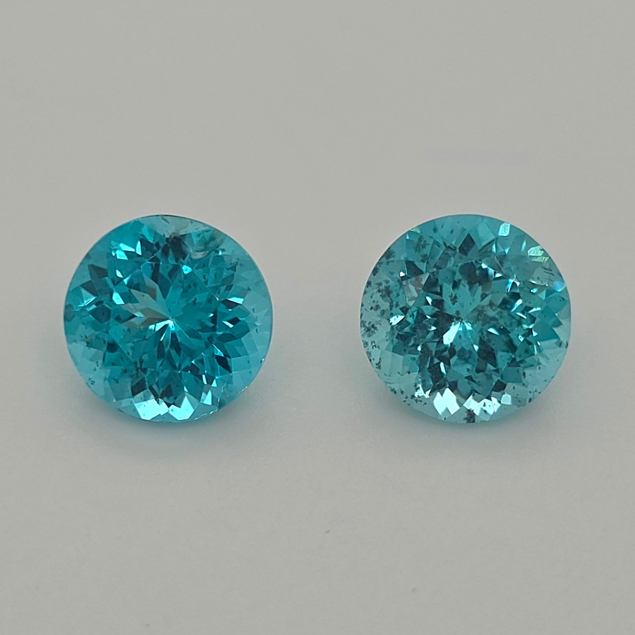Two round brilliant-cut blue gemstones, displayed side-by-side on a neutral light gray background.