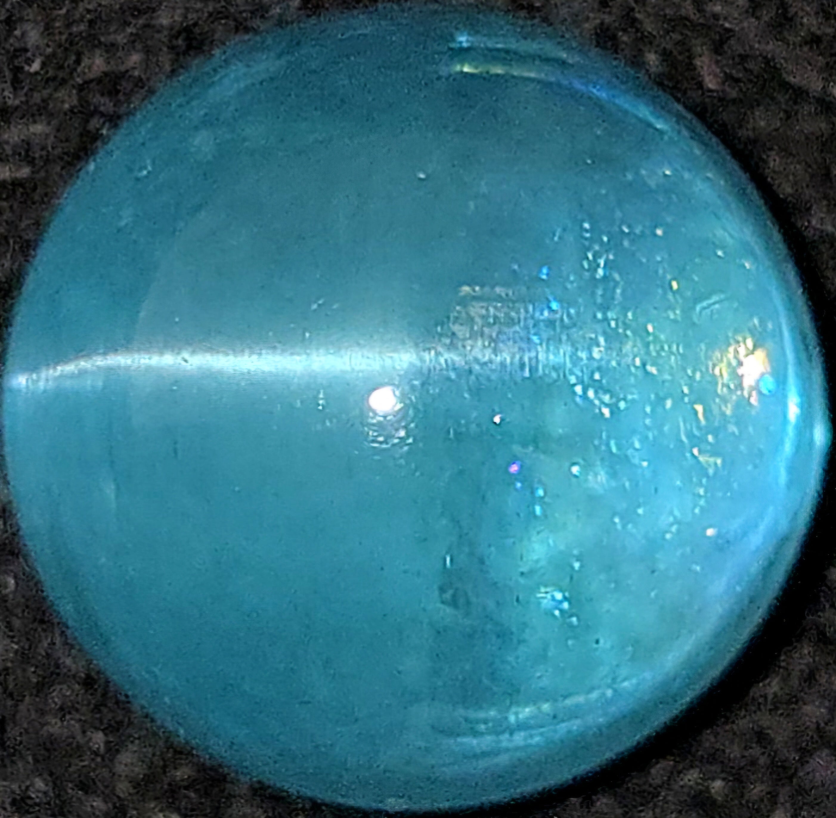 A round, polished turquoise-blue cabochon gemstone with a smooth, glossy surface and a bright horizontal highlight, resting on a dark textured background.