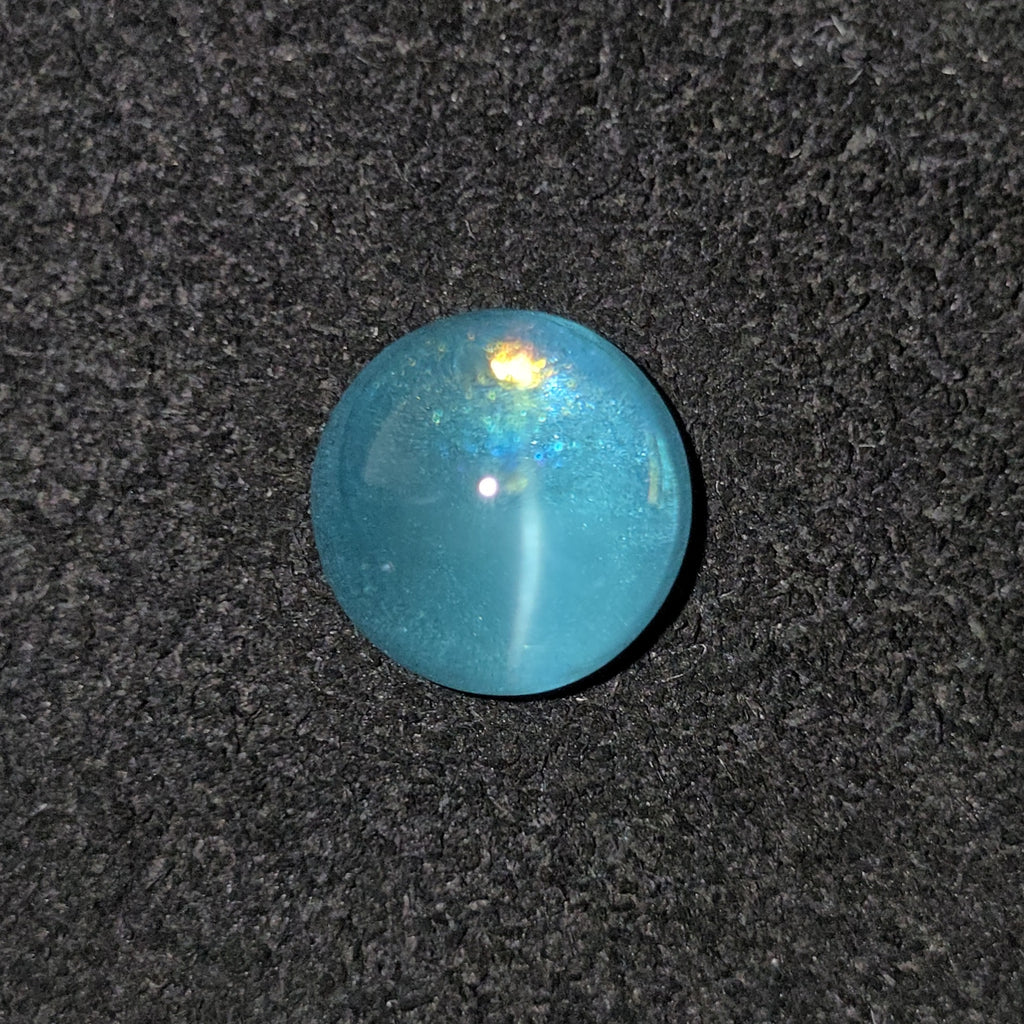 A round, polished turquoise-blue cabochon gemstone with a subtle iridescent sheen, displayed on a textured dark gray background.