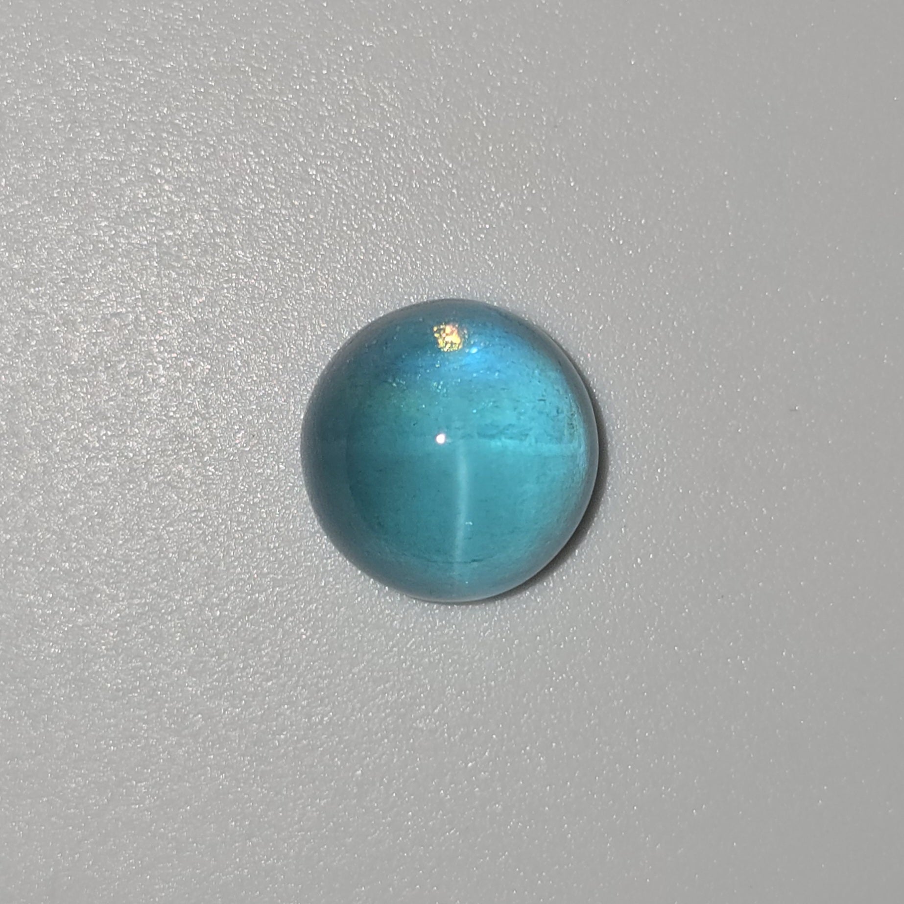 A single round, polished turquoise cabochon gemstone with a subtle cat's-eye reflection, displayed on a textured light gray background.