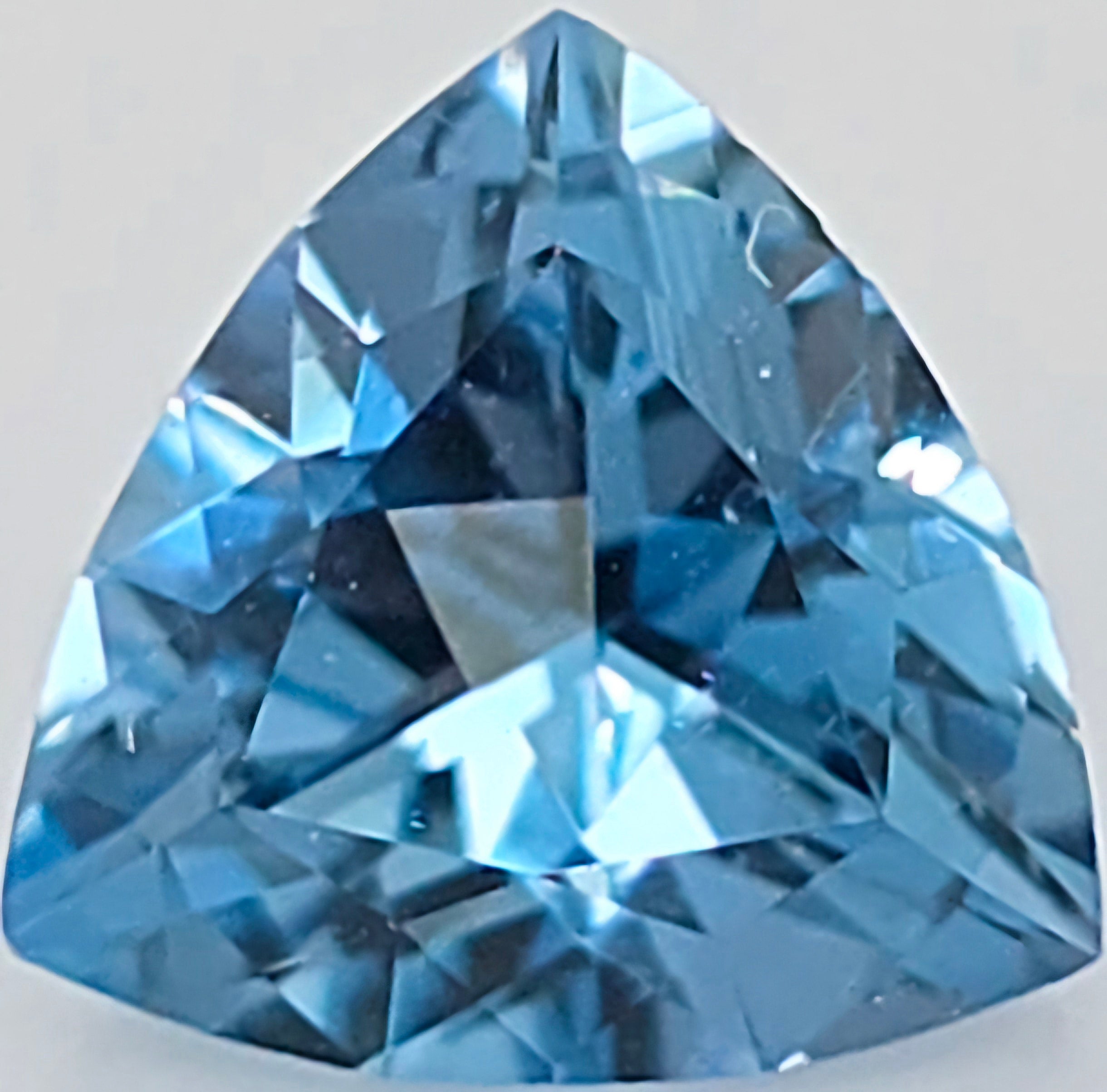A faceted triangular blue gemstone, shown in close-up against a light gray background.