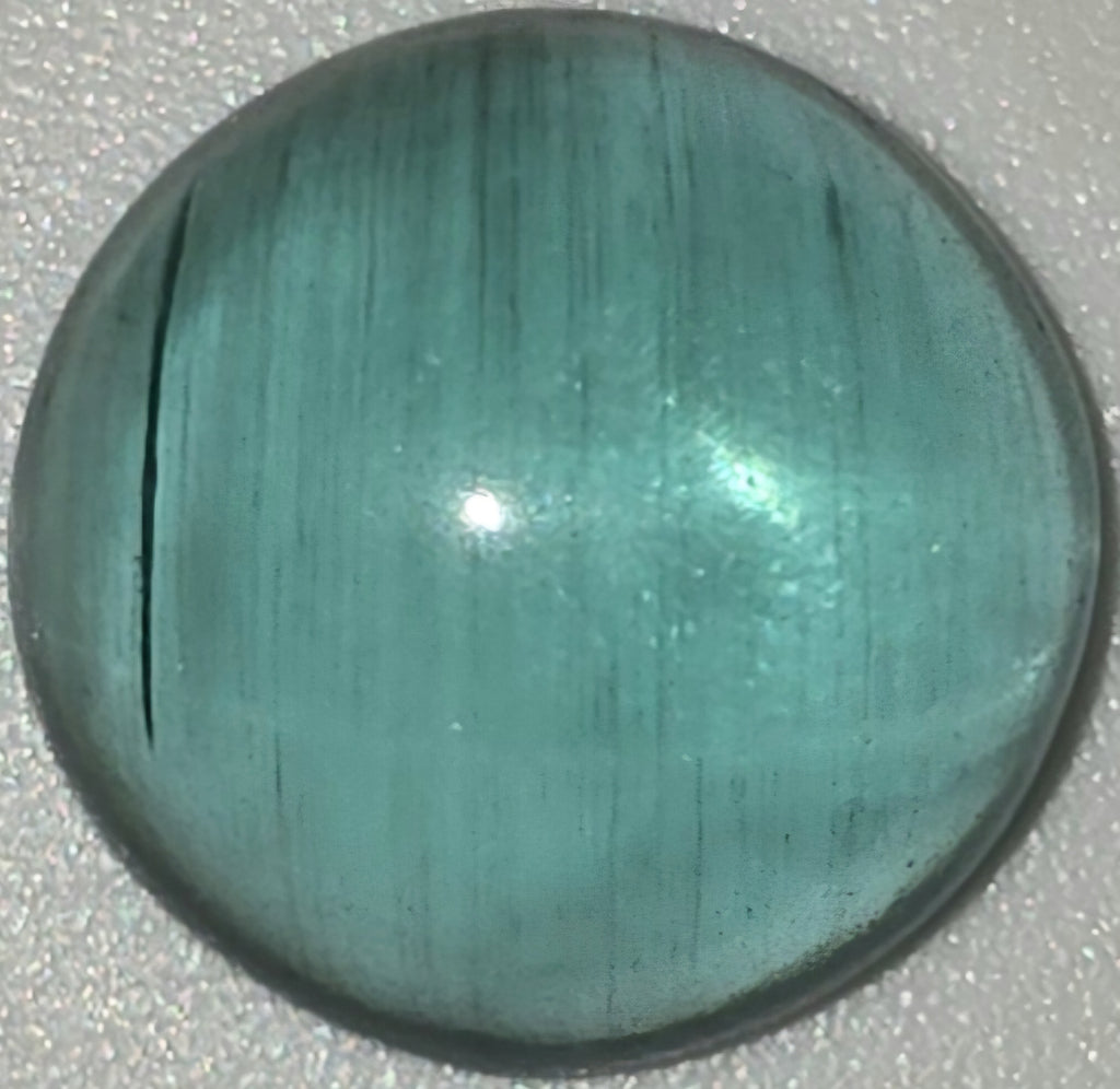 A polished round cabochon gemstone with a teal-green color and fine linear texture, shown on a textured light gray background.