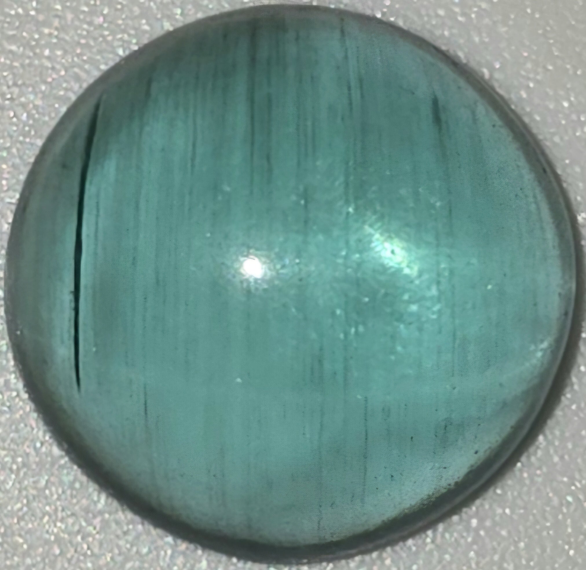 A polished round cabochon gemstone with a teal-green color and fine linear texture, shown on a textured light gray background.