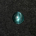 A round, dome-shaped bluish green gemstone with a bright white starburst reflection, resting on a dark textured fabric background.