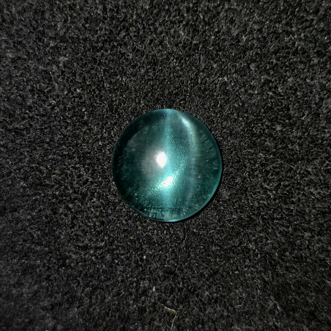 A round, dome-shaped bluish green gemstone with a bright white starburst reflection, resting on a dark textured fabric background.