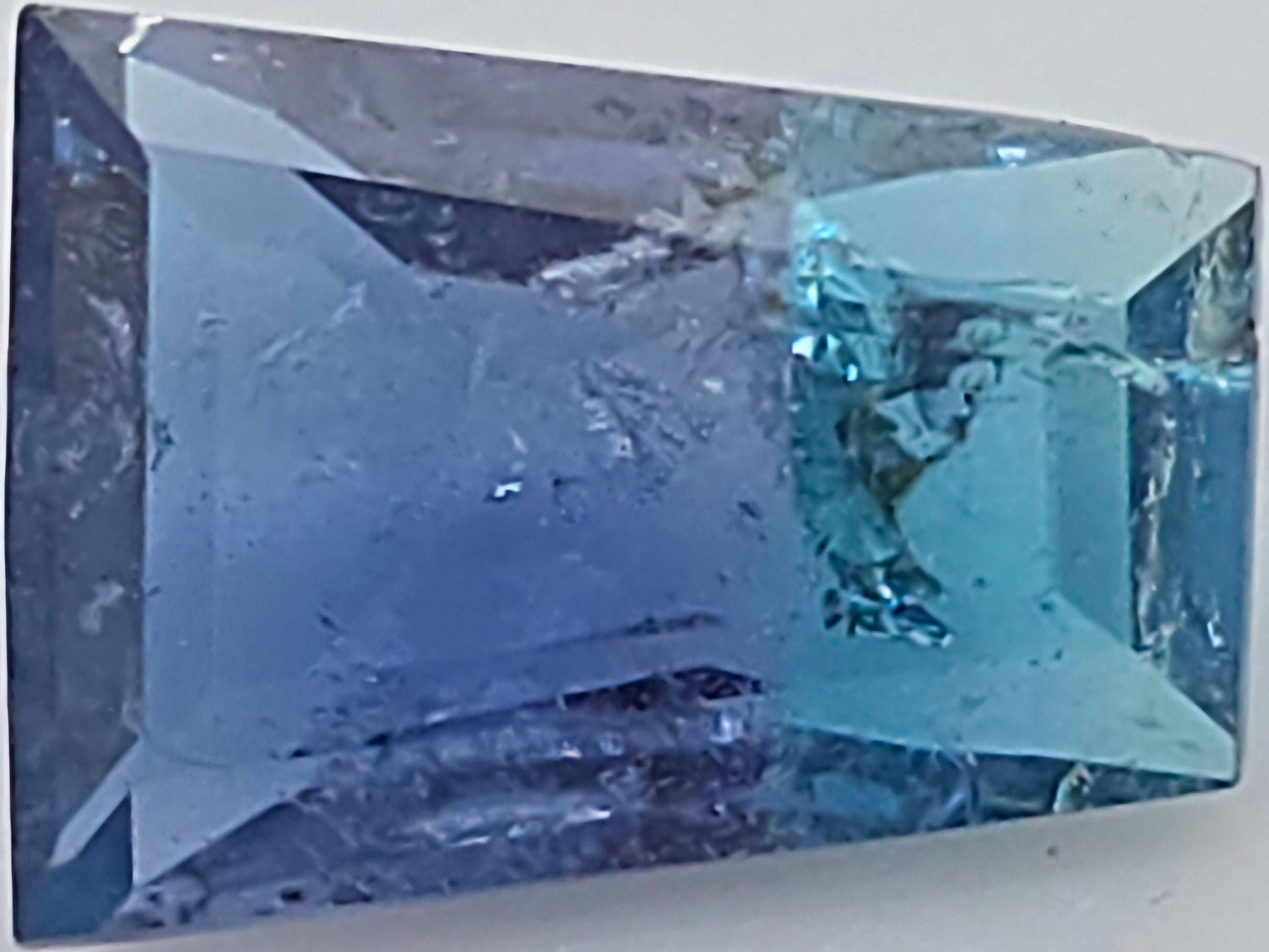 A trapezoid shaped gemstone with a blue to turquoise gradient, displayed on a neutral light gray background.