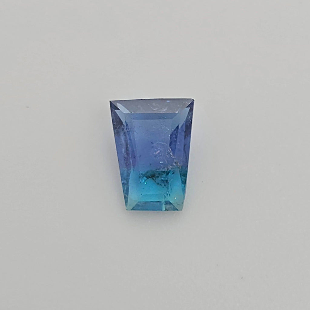 A trapezoid shaped gemstone with a blue to turquoise gradient, displayed on a neutral light gray background.