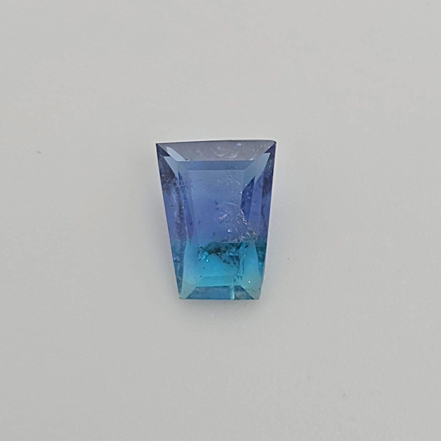 A trapezoid shaped gemstone with a blue to turquoise gradient, displayed on a neutral light gray background.