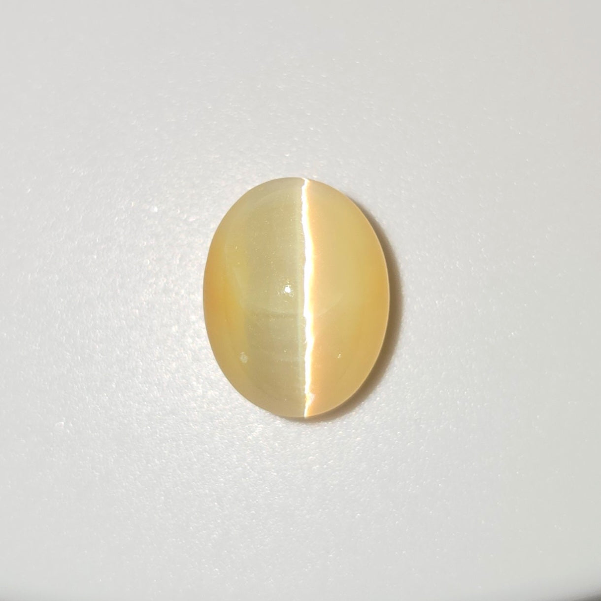 An oval cat's eye gemstone with a pale yellow body and a distinct white split reflection, displayed on a clean white background.