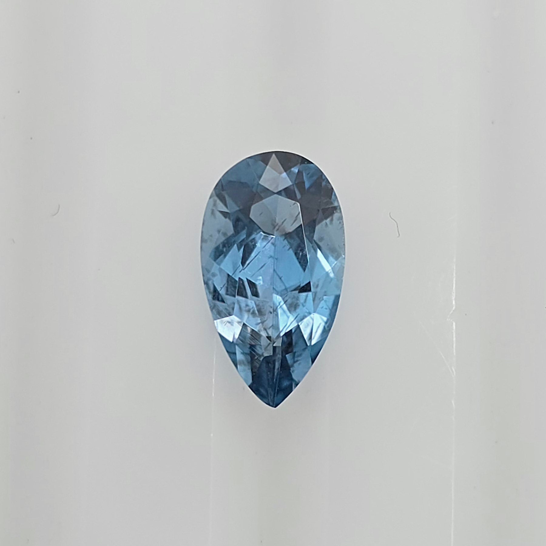 A single pear-shaped blue gemstone with a faceted cut, centered on a plain light gray background.