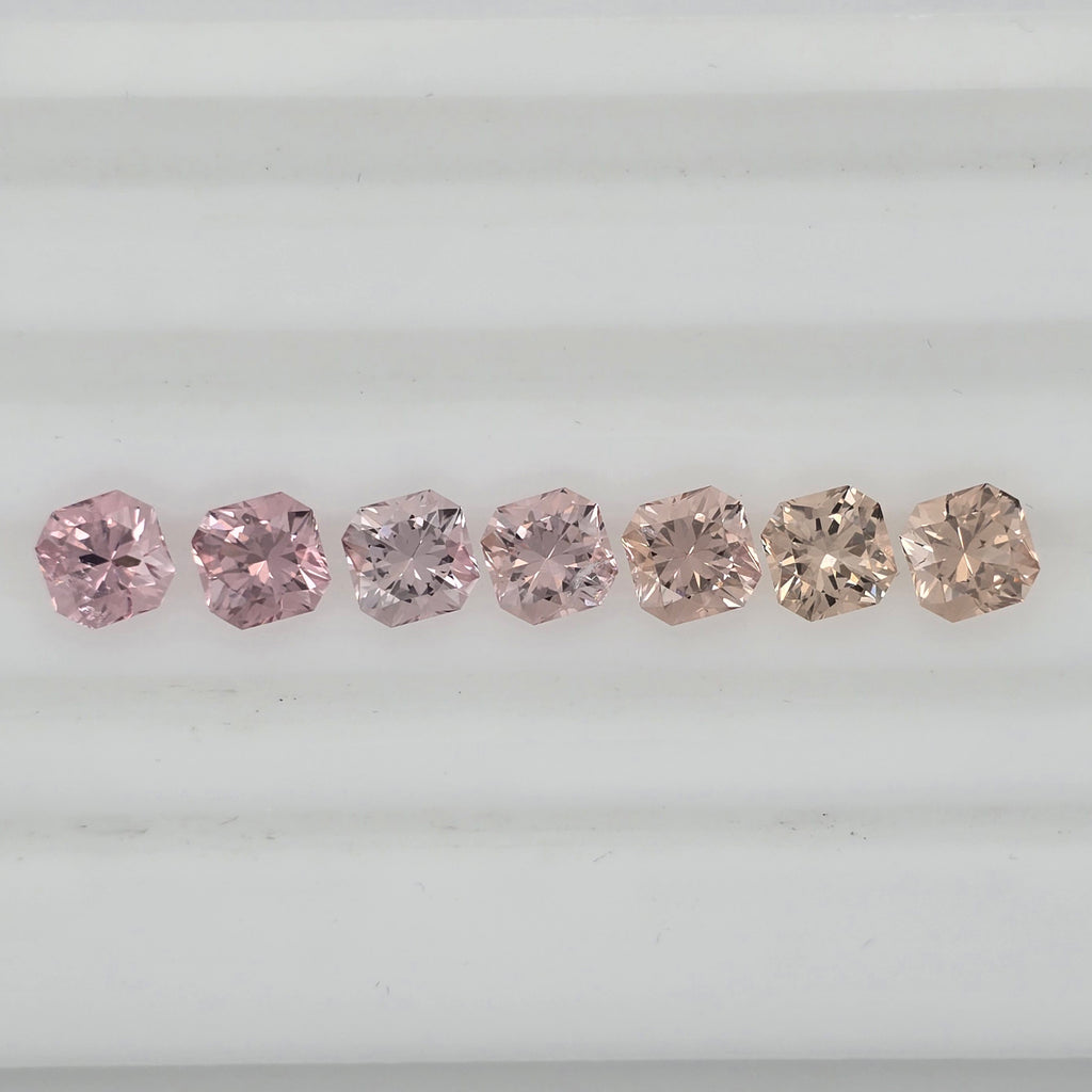 Seven square-cut pink gemstones arranged in a row on a white background, showing varying shades from pink to pinkish orange