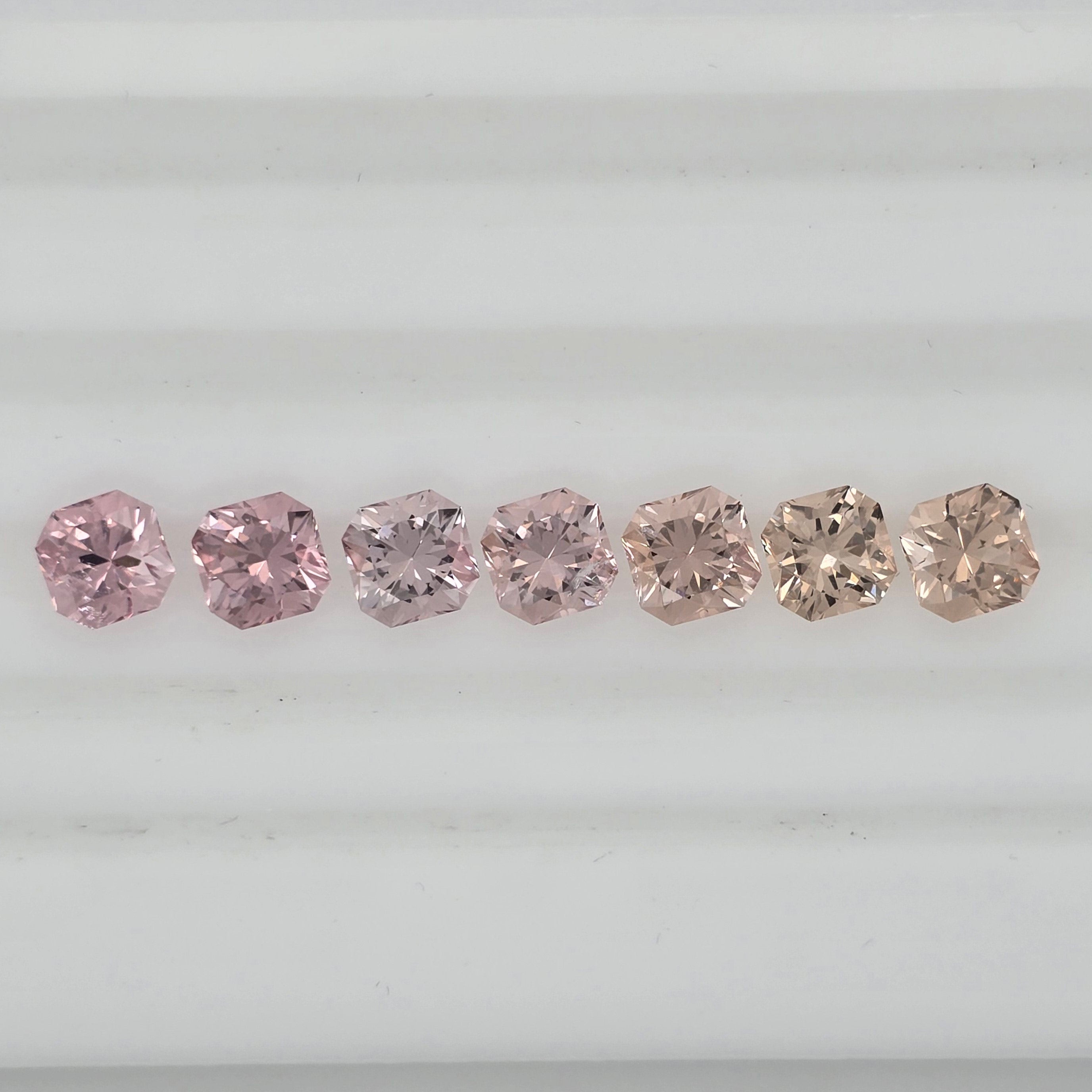 Seven square-cut pink gemstones arranged in a row on a white background, showing varying shades from pink to pinkish orange