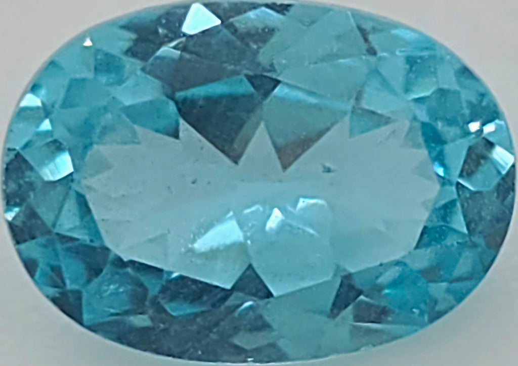 A faceted oval-cut greenish blue gemstone shown against a plain light gray background.