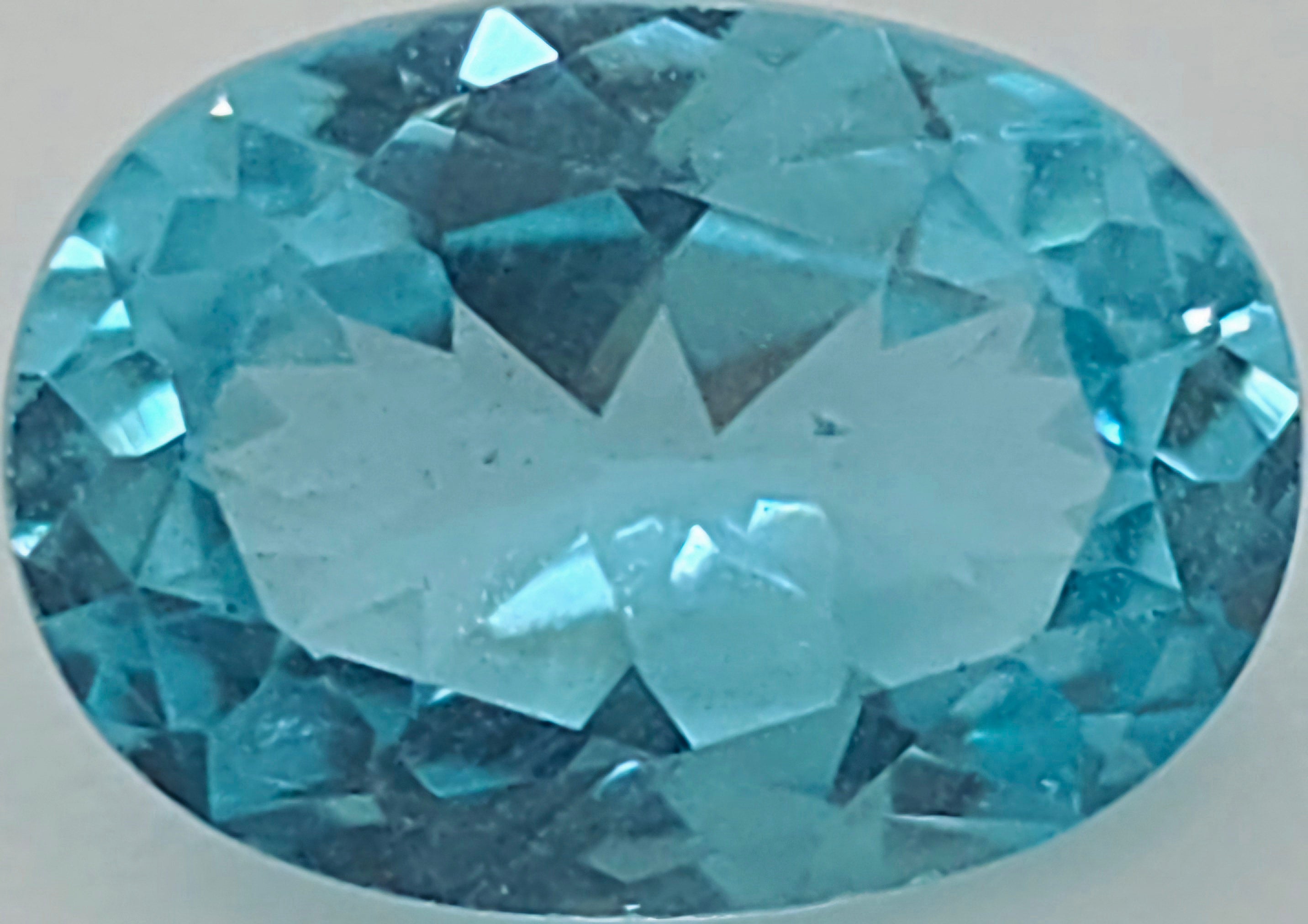 A faceted oval-cut greenish blue gemstone shown against a plain light gray background.