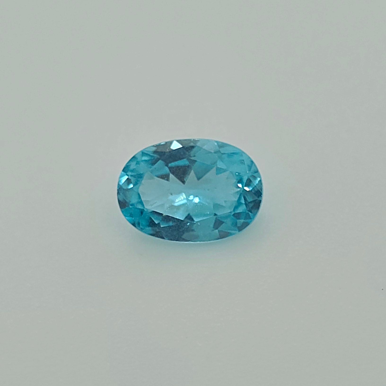 A faceted oval-cut greenish blue gemstone centered on a plain light gray background.