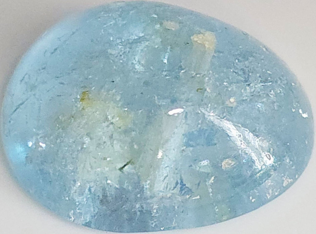 A polished oval-shaped light blue gemstone with natural inclusions and a glossy finish, shown on a plain light background.