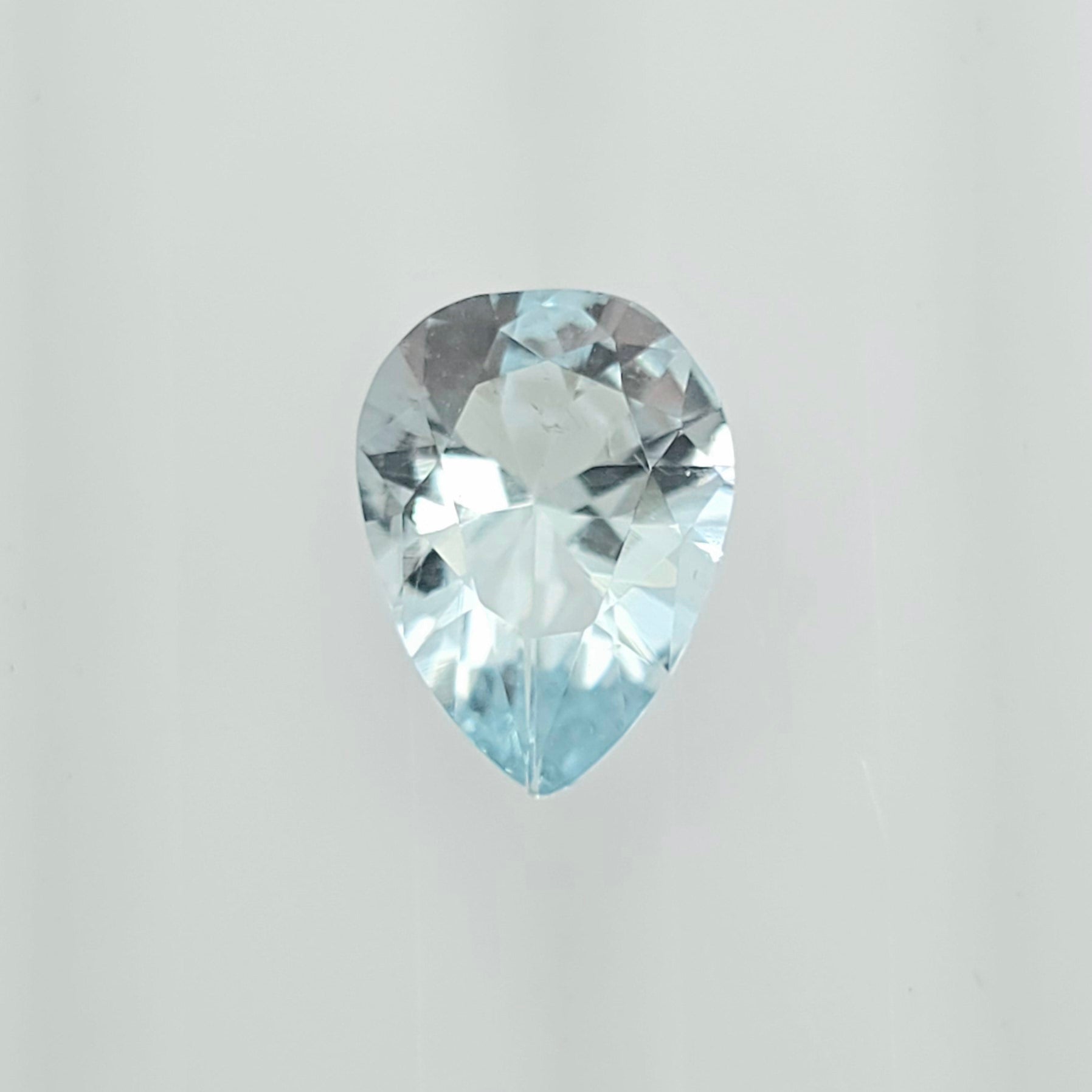 A single pear-cut light blue gemstone with a faceted, reflective surface, centered against a plain light gray background.