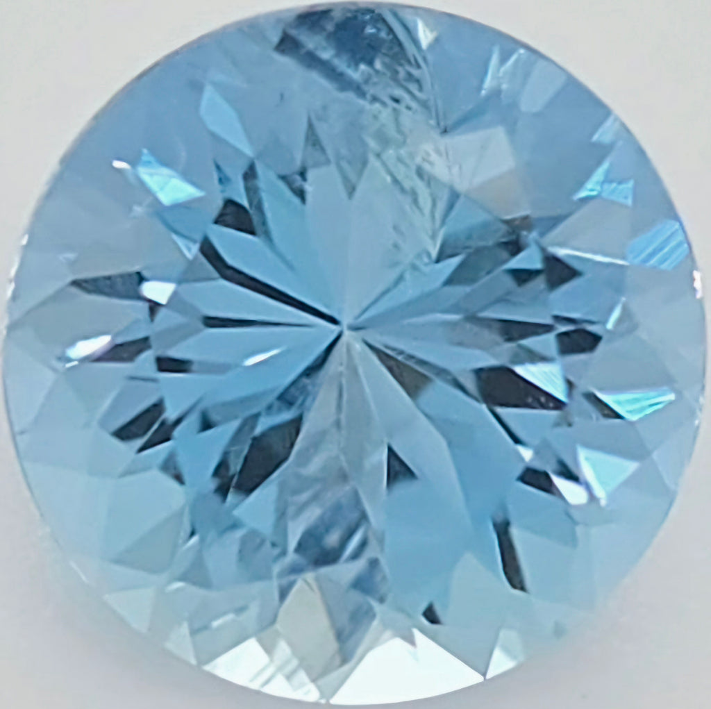 A round brilliant-cut light blue gemstone, showing detailed faceted reflections on a plain light background.